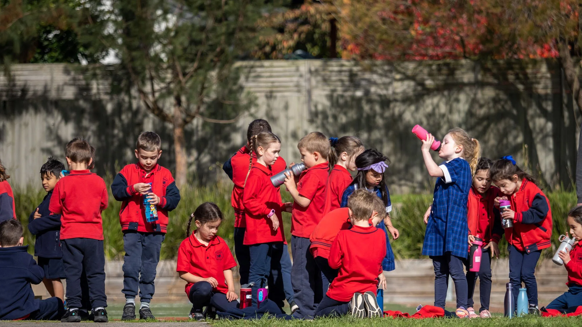 Children in red and blue uniforms gathering outside, some drinking water and others standing or sitting on the grass, with trees and a fence in the background.