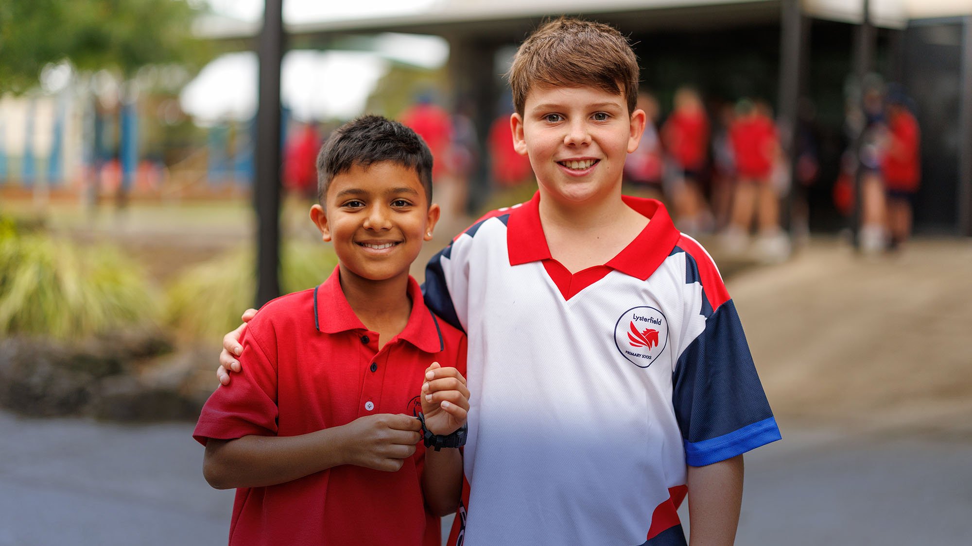 Two boys in school uniforms smiling, with students in red uniforms in the background. One boy is wearing a red polo shirt, and the other is in a white, red, and blue sports shirt with a logo that reads "Lysterfield Primary School."