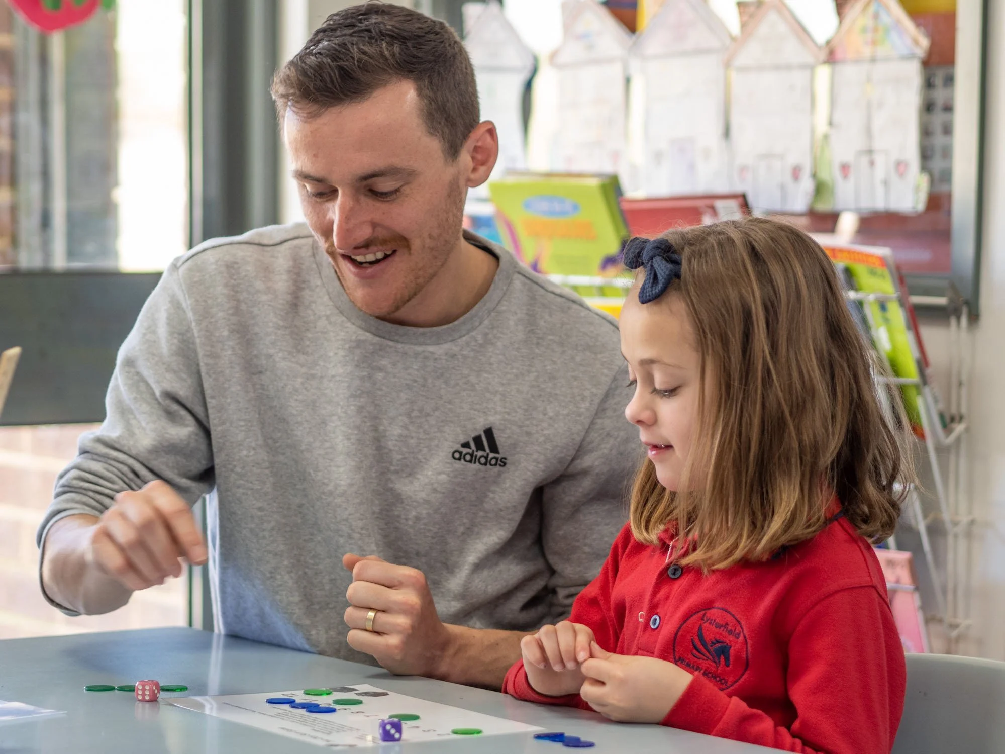 A man and a young girl playing a board game with colorful game pieces, coins, and dice at a table in a classroom or playroom.