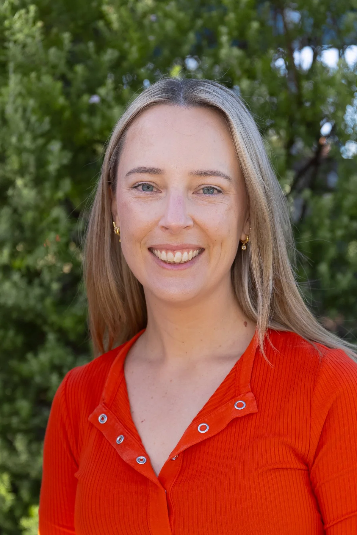 A woman with blonde hair, light skin, and blue eyes, smiling outdoors in front of green trees, wearing a bright orange top and gold earrings.