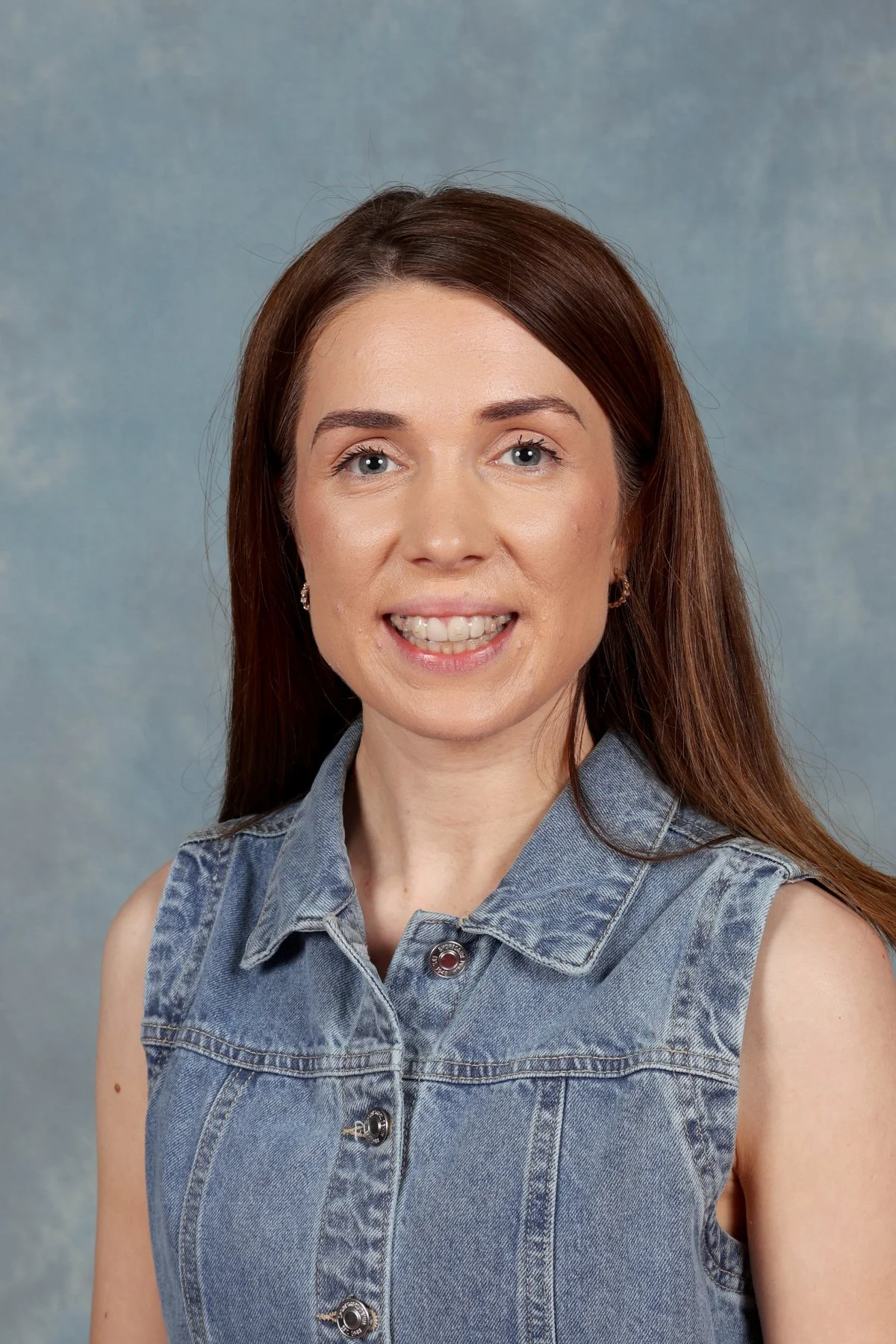 A woman with long red hair wearing a sleeveless denim vest, smiling, against a plain background.