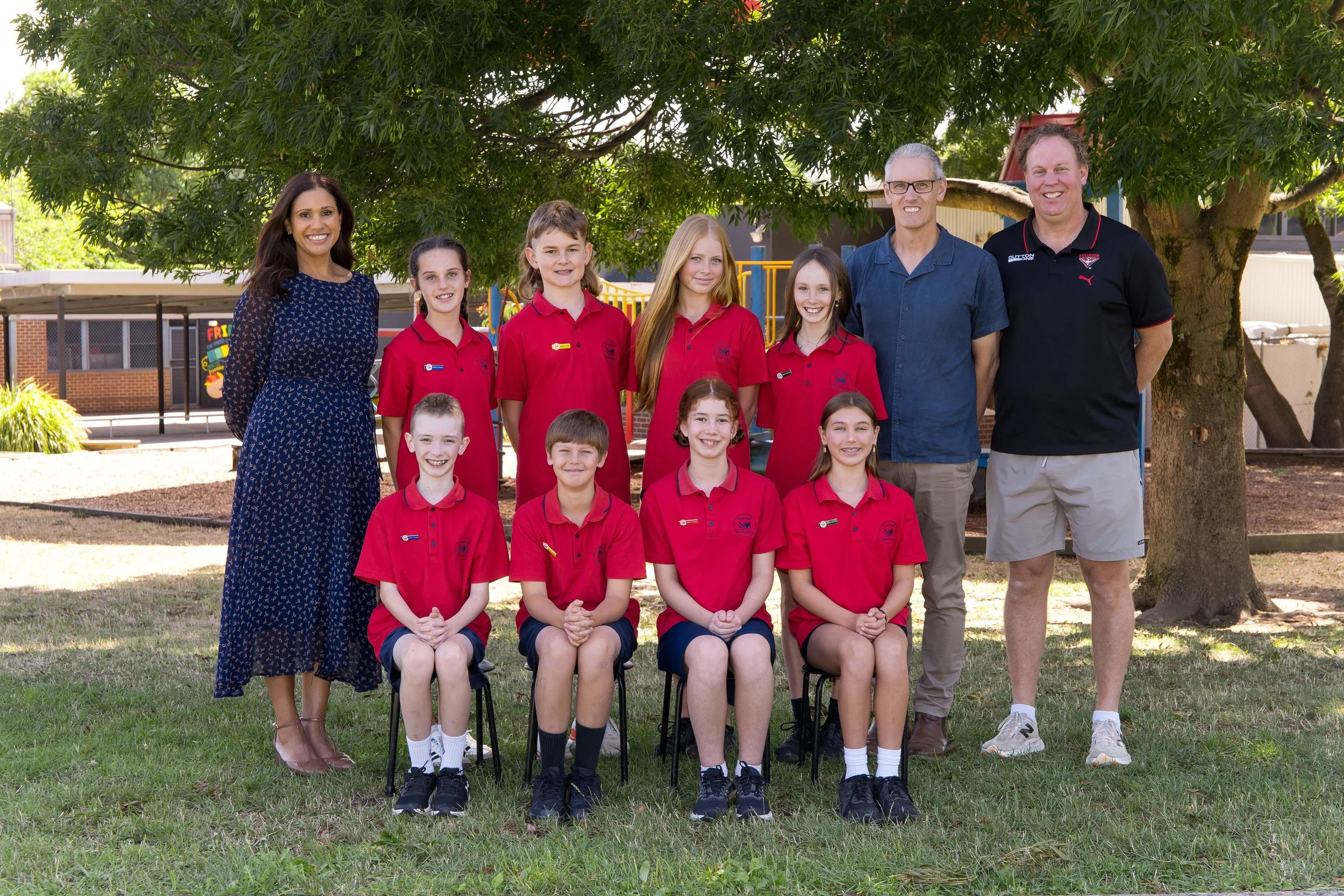 A group of children and adults posing outdoors on a grassy area with a tree and playground in the background. The children are wearing red uniforms and sitting on small chairs, while the adults stand behind them. The group is smiling.