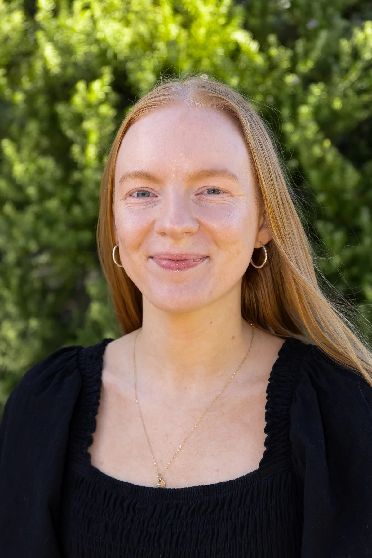 A young woman with red hair, freckles, and hoop earrings, smiling outdoors with a blurred green leafy background.