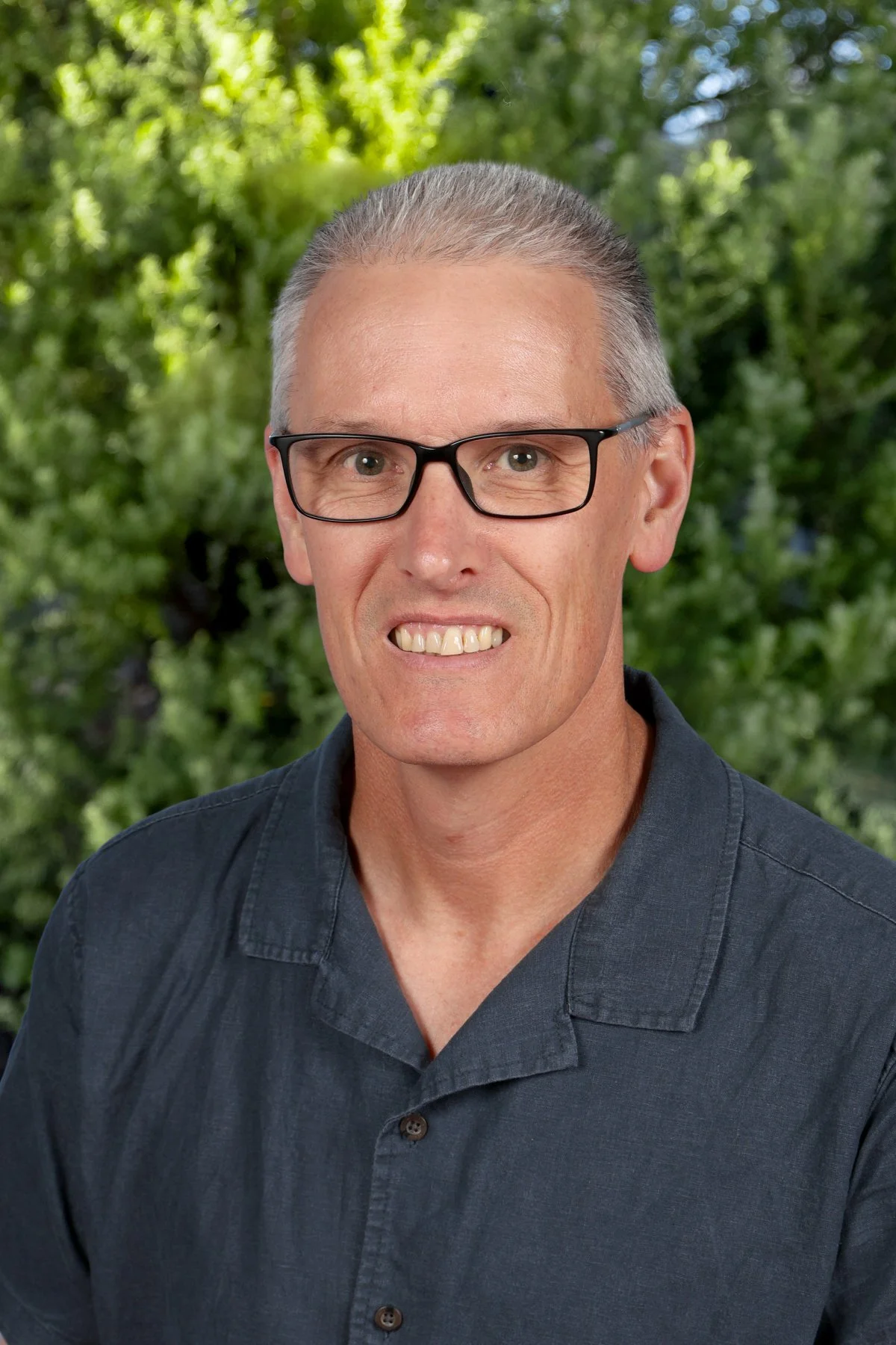 A middle-aged man with short gray hair, glasses, and a black button-up shirt is smiling slightly, with a background of green trees and foliage.