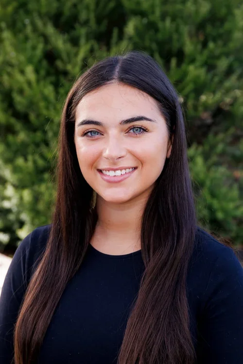 Young woman with long dark hair, smiling, wearing a black top outdoors with green foliage in the background.