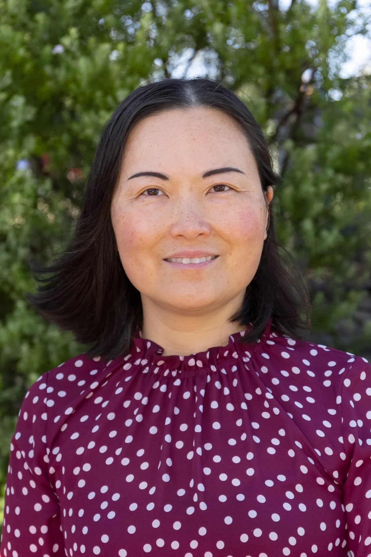 A woman with shoulder-length dark hair smiling outdoors in front of green trees, wearing a burgundy blouse with white polka dots.