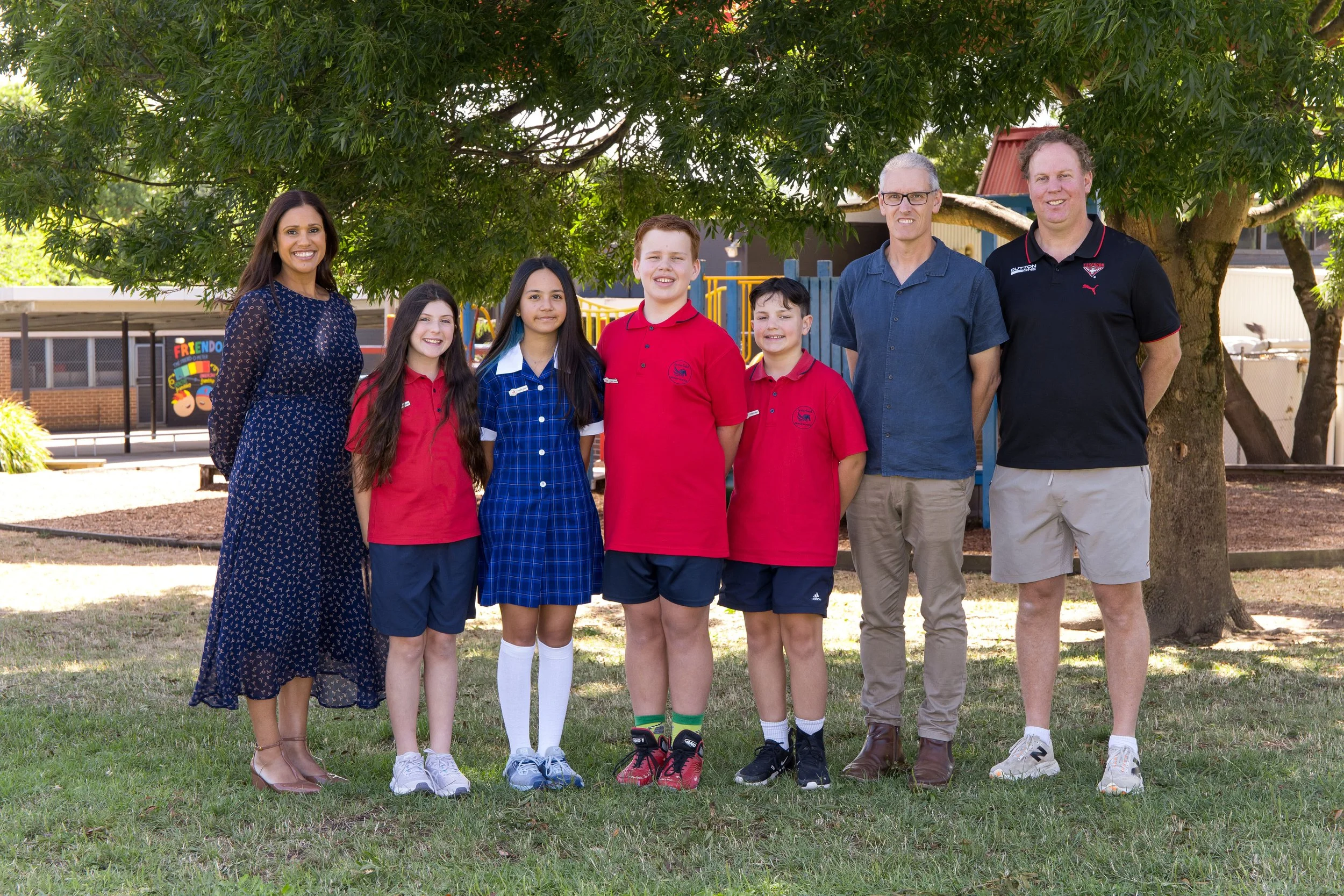 Group of seven people, three adults and four children, standing outdoors on grass under a large tree. In the background, there is a playground and a fence.
