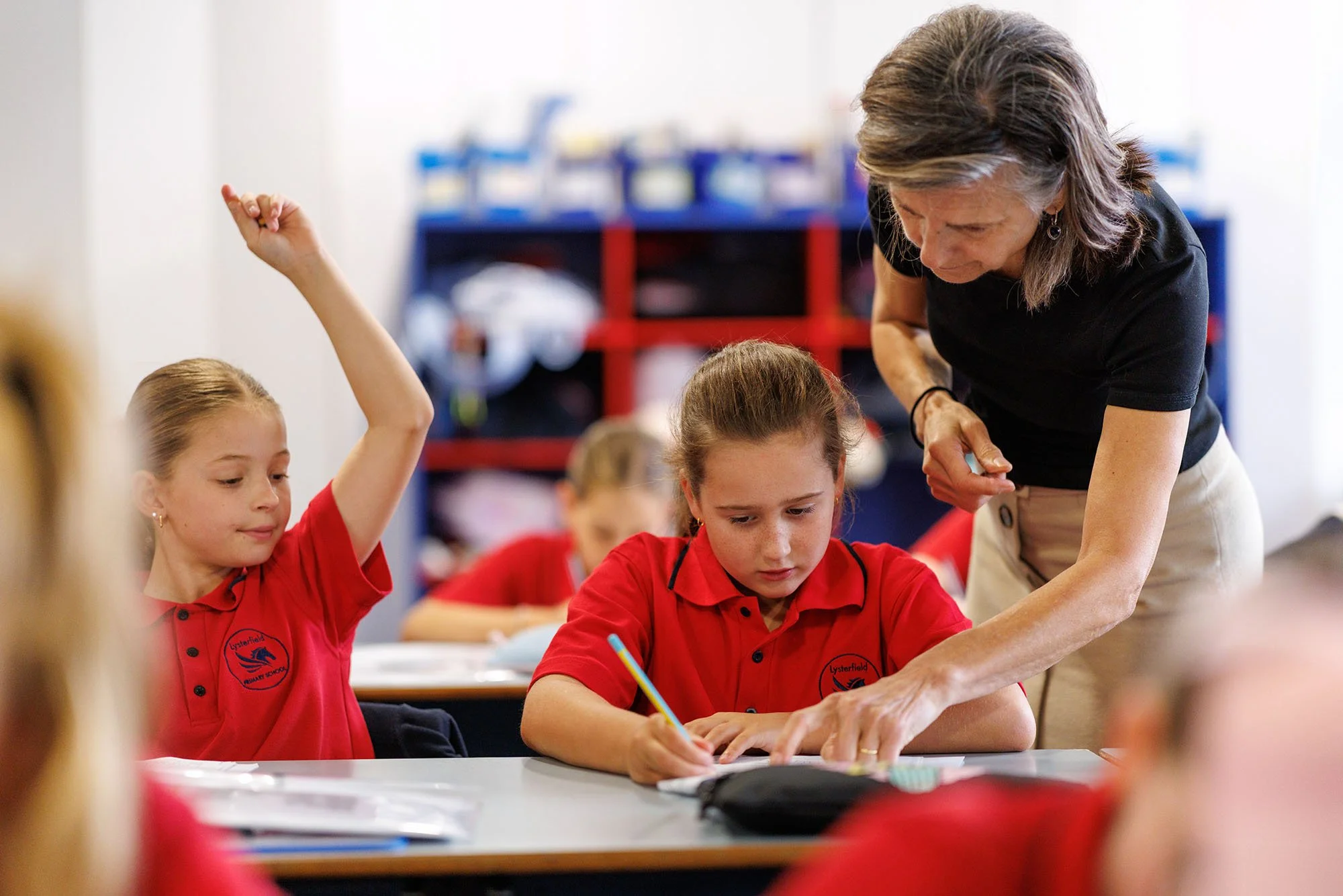 A female teacher helping a girl in a red school uniform with her work in a classroom. Another girl in a red uniform has her hand raised.