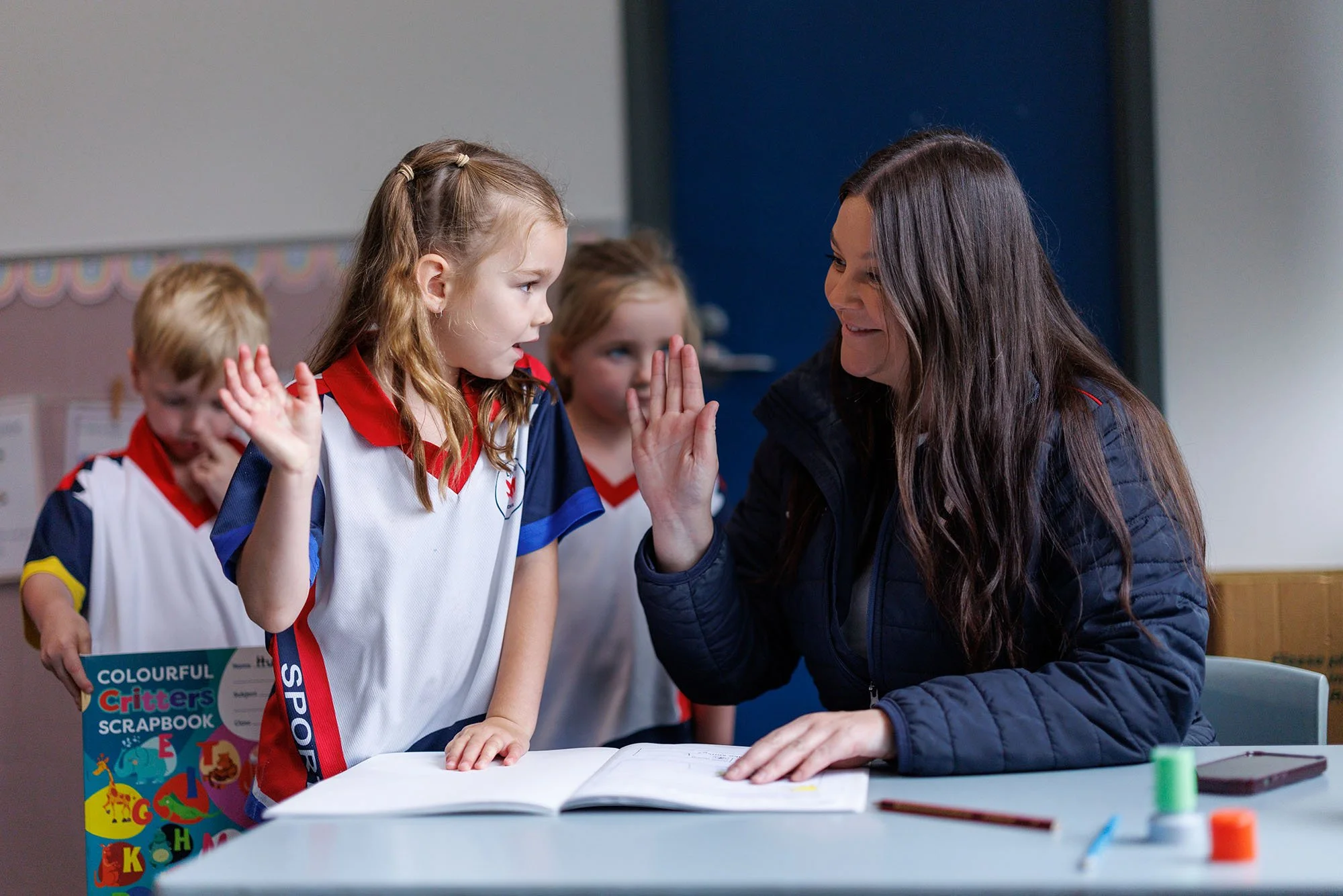 A teacher giving a high-five to a young girl in a classroom with other children.