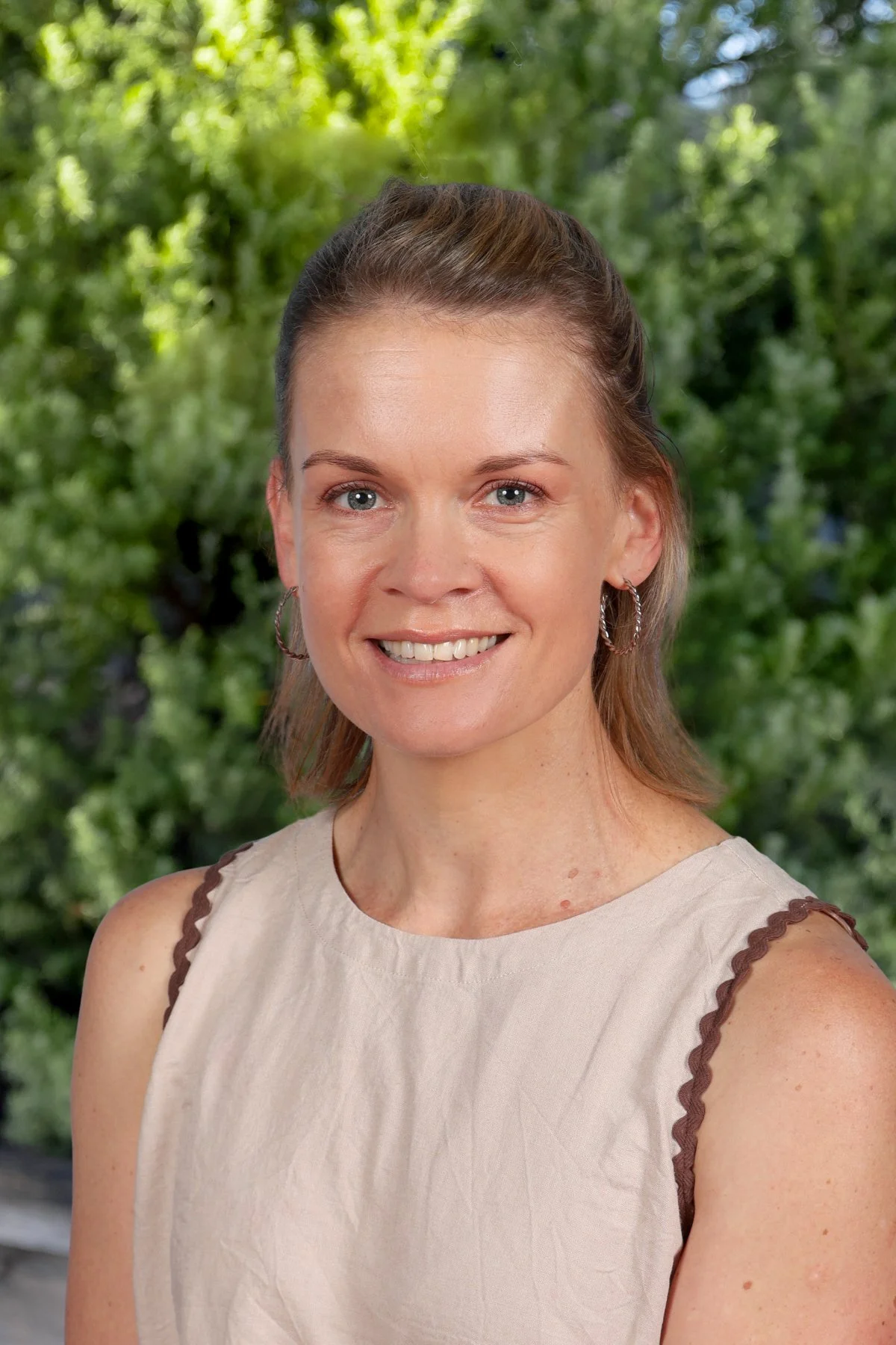 A woman with light brown hair tied in a ponytail, wearing hoop earrings and a sleeveless beige top with brown scalloped details on the shoulders, standing outdoors in front of green trees and foliage, smiling at the camera.