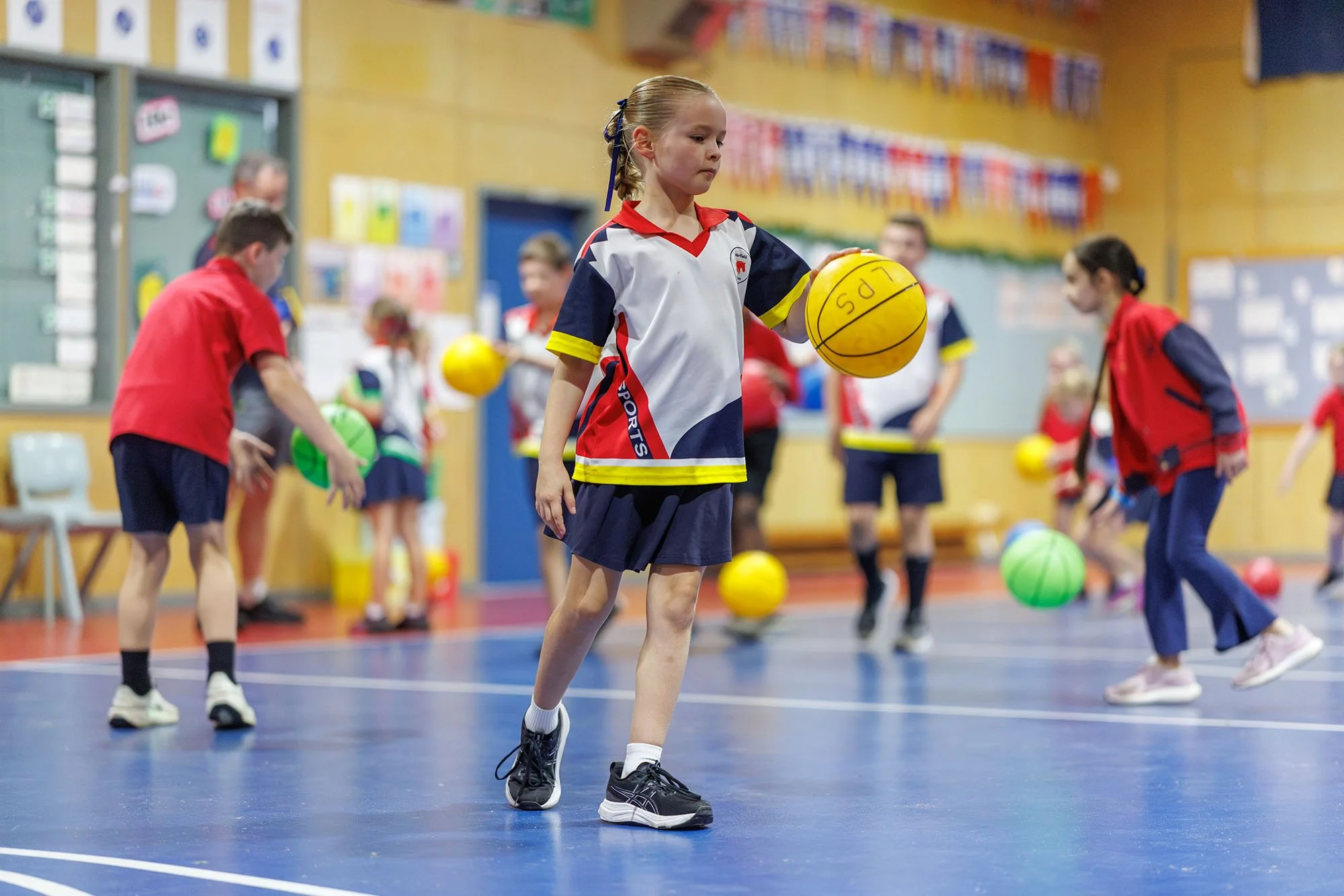 Young girl holding a yellow sports ball during a physical education class in a school gymnasium with other children playing with various sports balls in the background.