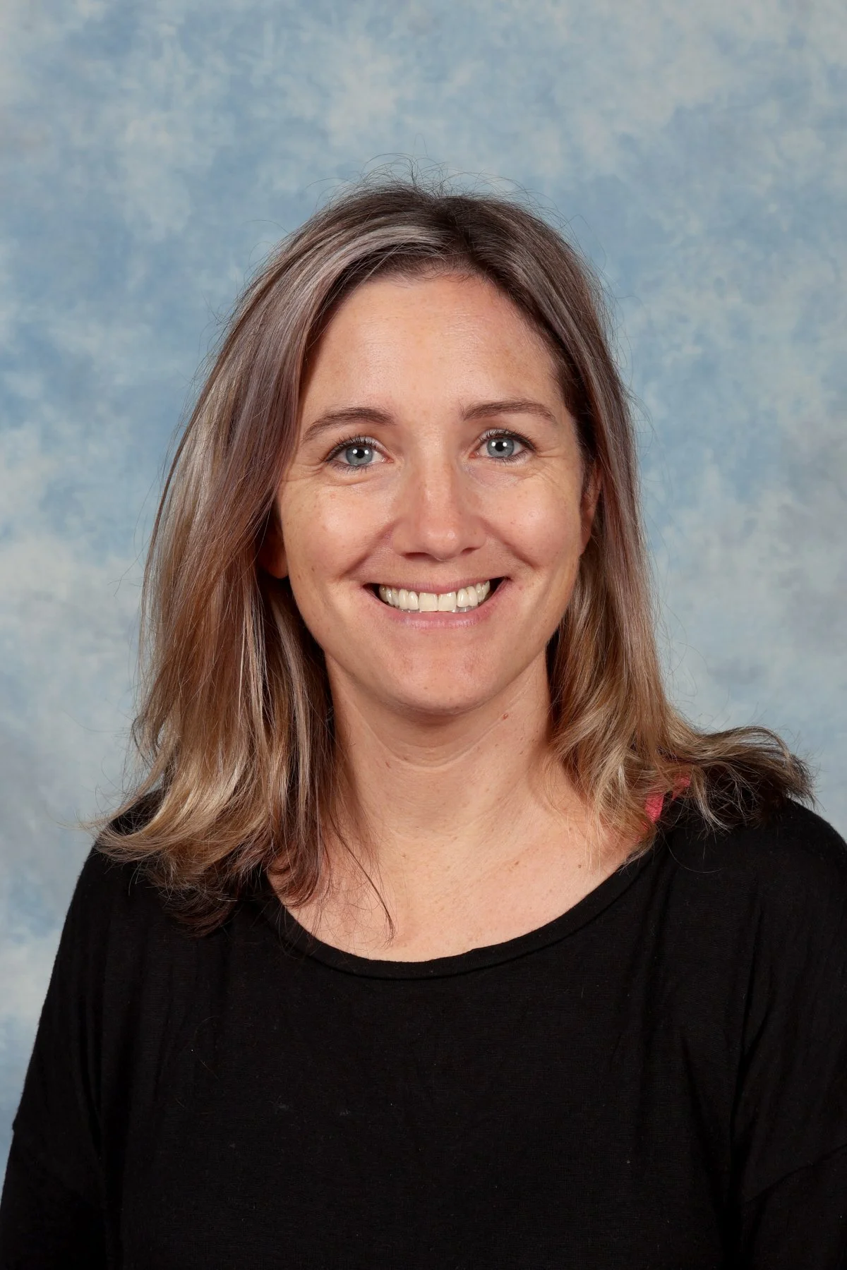 A woman with shoulder-length light brown hair and blue eyes smiling at the camera, wearing a black top against a blue sky background.