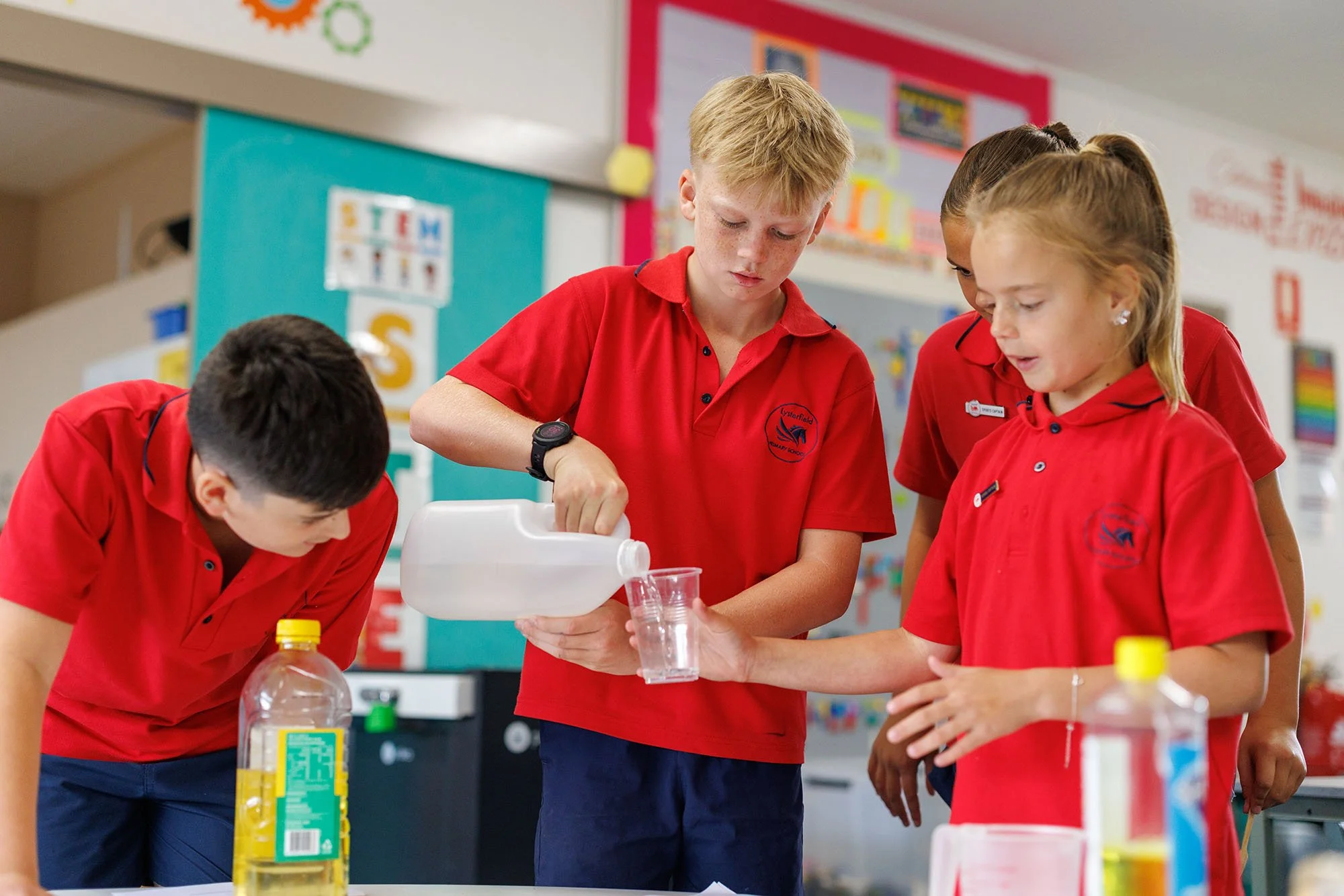 Four students wearing red shirts are conducting a science experiment with liquids in a classroom.