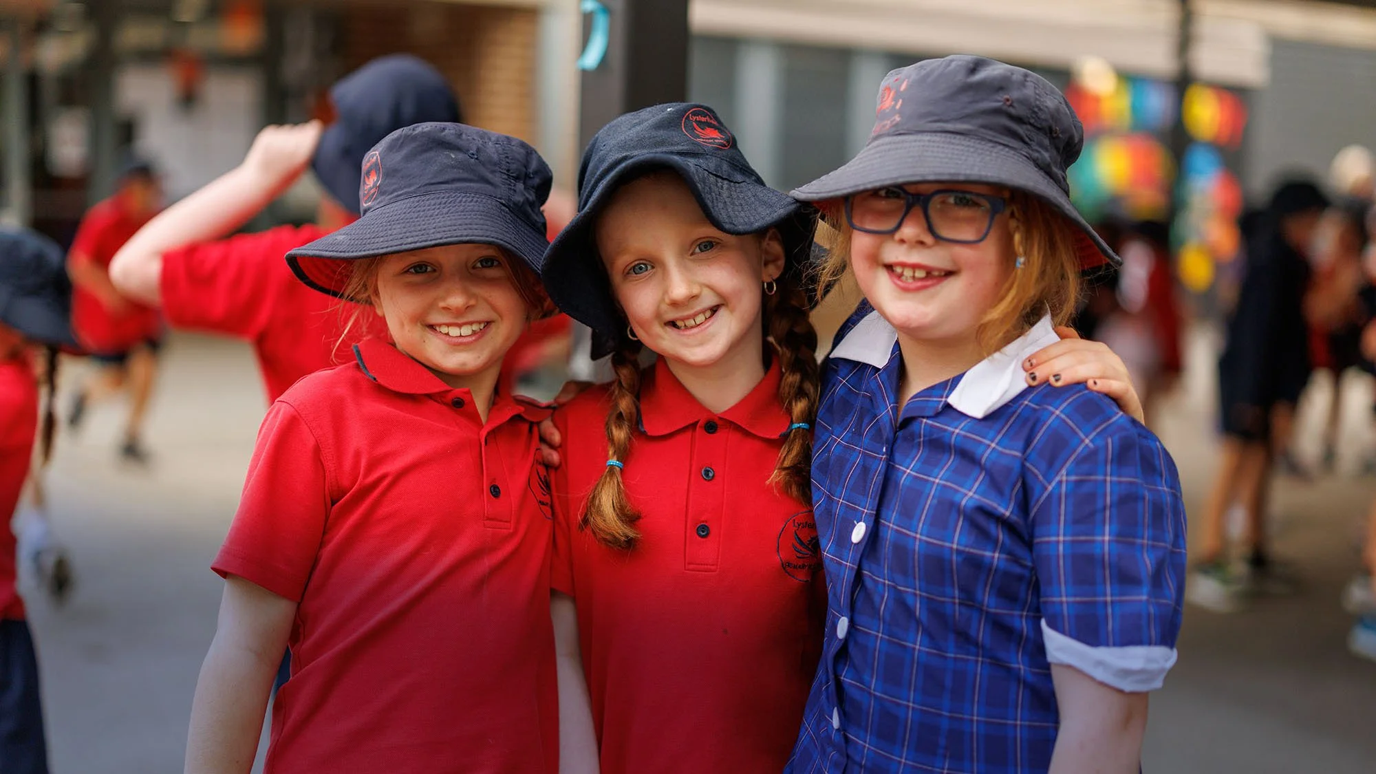 Three school-aged girls wearing hats and school uniforms are smiling and standing together outdoors in the school yard.