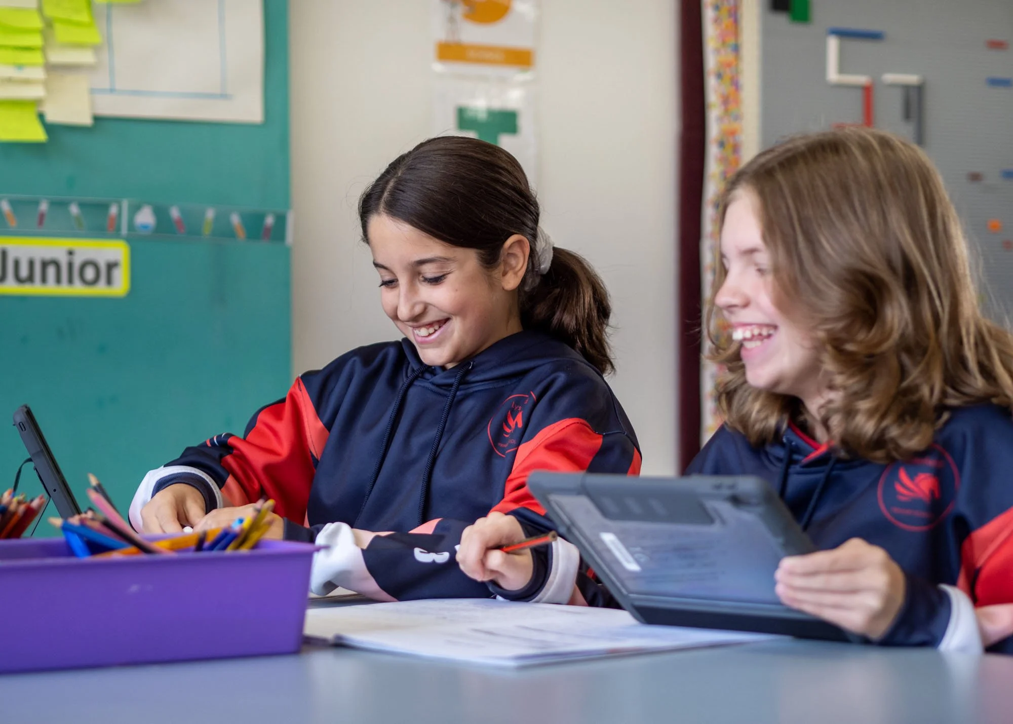 Two young girls in matching navy and red school uniforms sit at a desk, smiling and engaged with tablets and pencils, in a classroom setting.