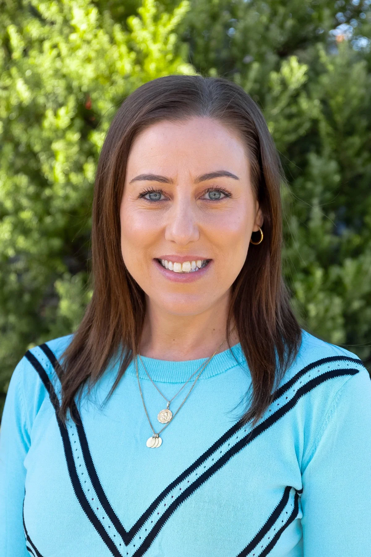 A young woman with long brown hair, blue eyes, and a warm smile standing outdoors with bright green trees in the background. She is wearing a light blue top with black accents, gold hoop earrings, and layered necklaces with gold charms.