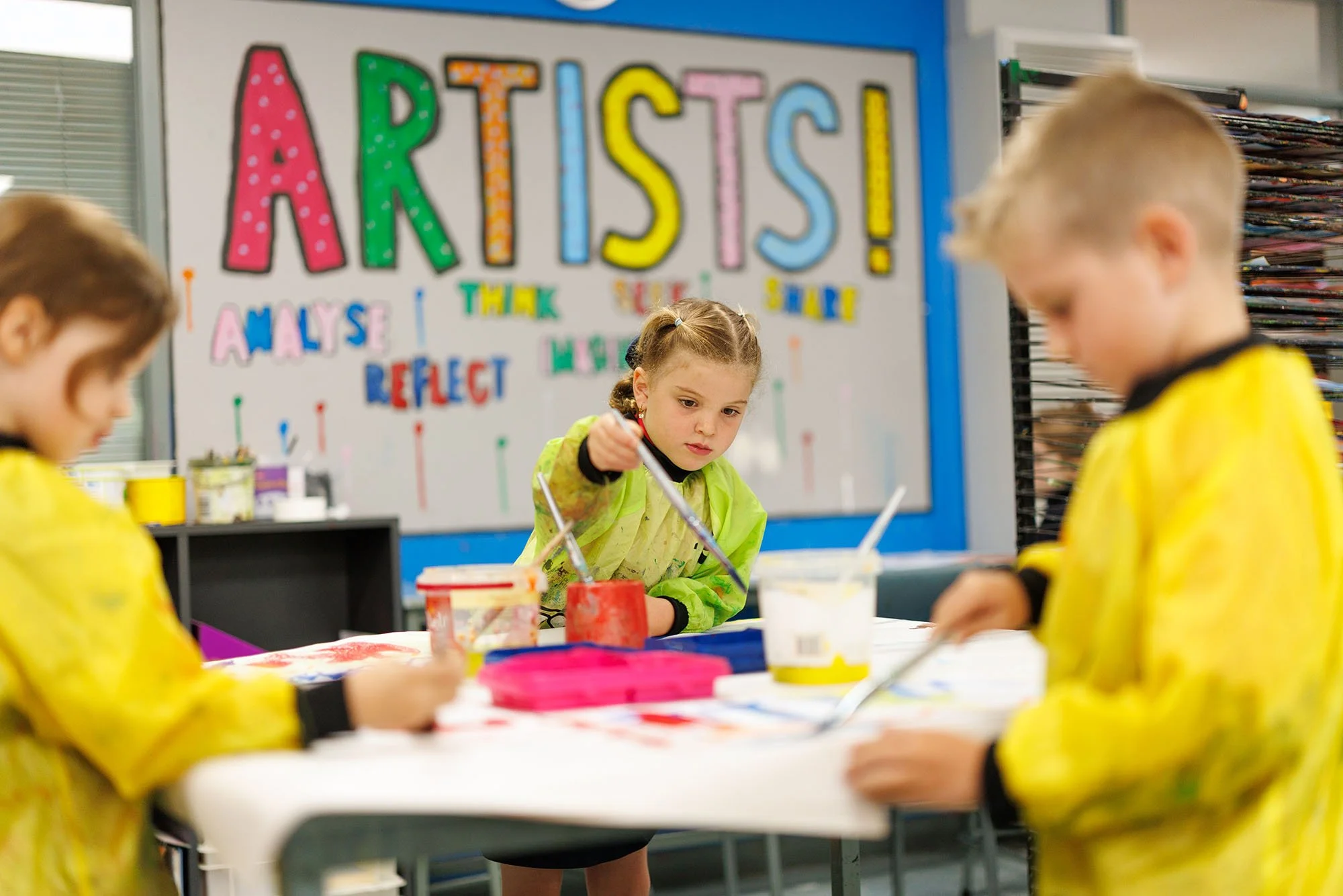 Children painting at a table in an art classroom with a colorful 'ARTISTS!' sign on the whiteboard in the background.