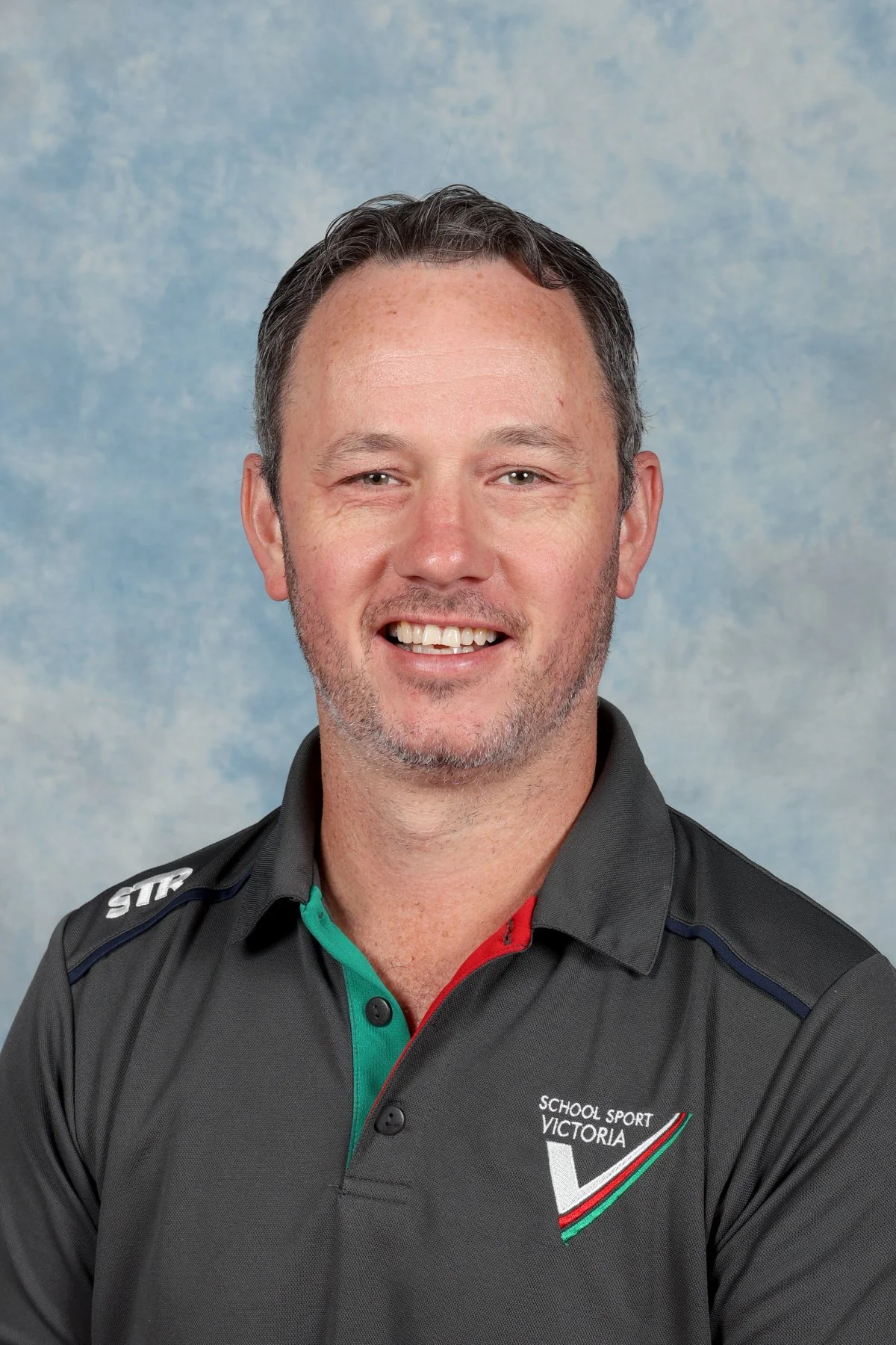 A smiling man with short dark hair and a beard, wearing a black sports polo shirt with a logo that reads 'School Sport Victoria' and a background of light blue clouds.