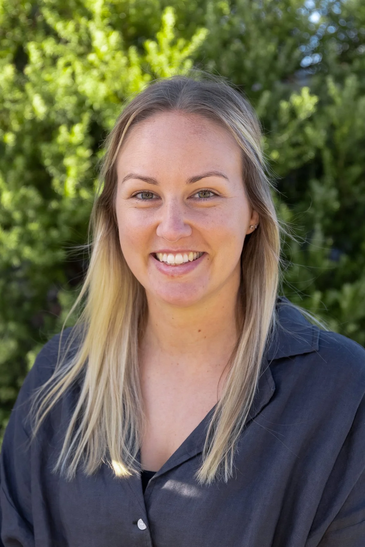Young woman with blonde hair, smiling outdoors with green trees in the background.