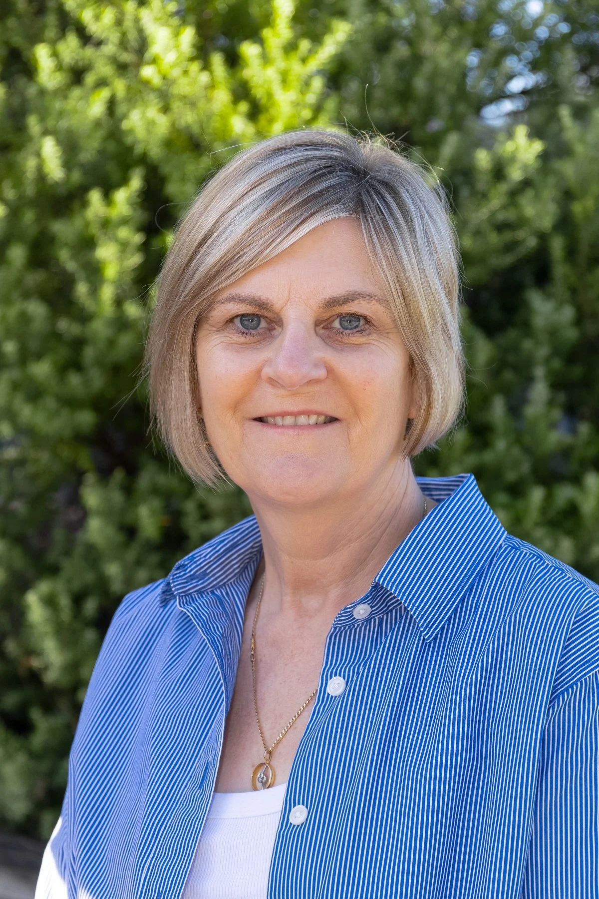 A woman with blonde hair and blue eyes smiling outdoors, wearing a blue striped shirt and a gold necklace with a pendant.