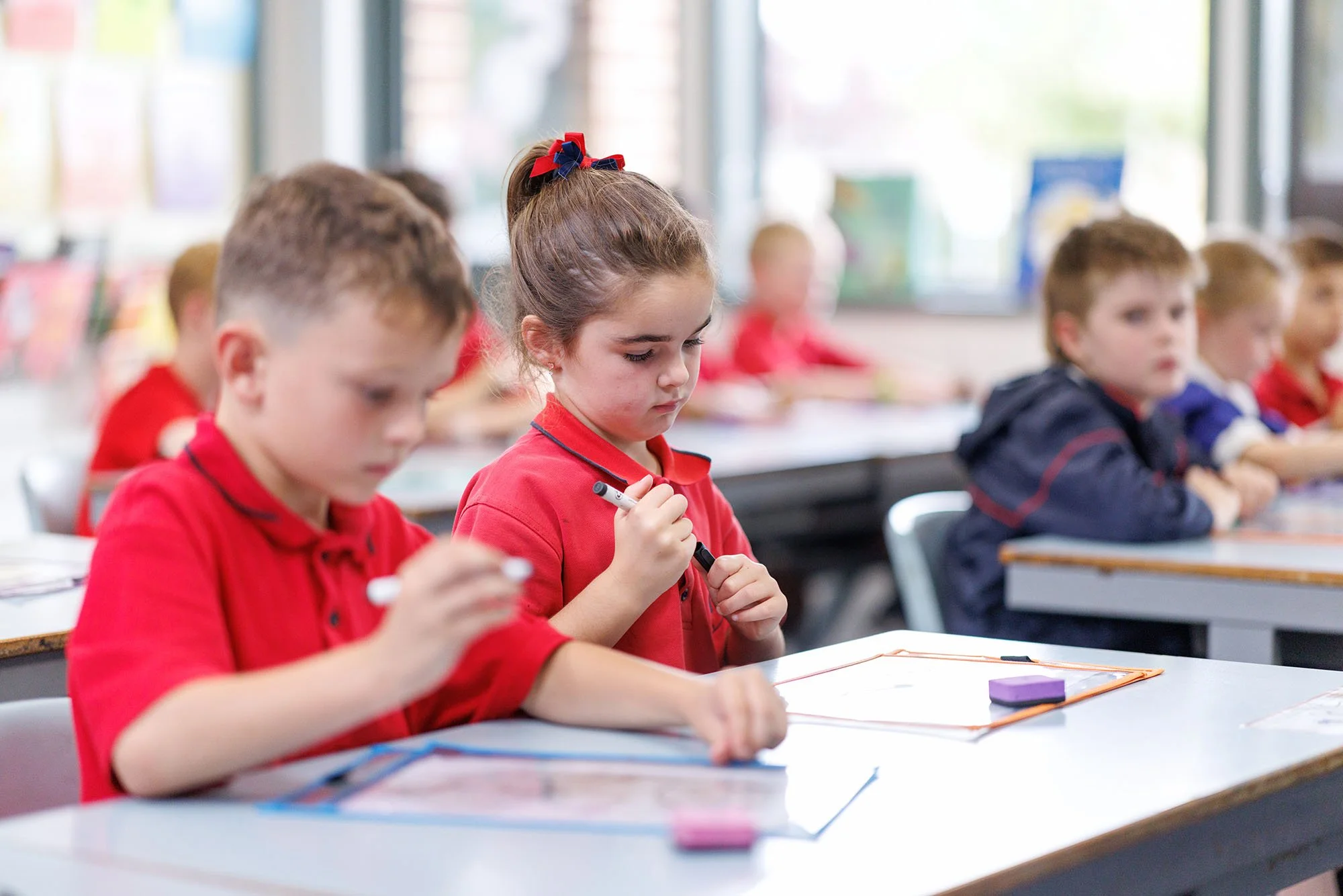 School children sitting at desks in a classroom, writing and reading, with a girl in the center wearing a red shirt and a red, black, and blue hair bow.