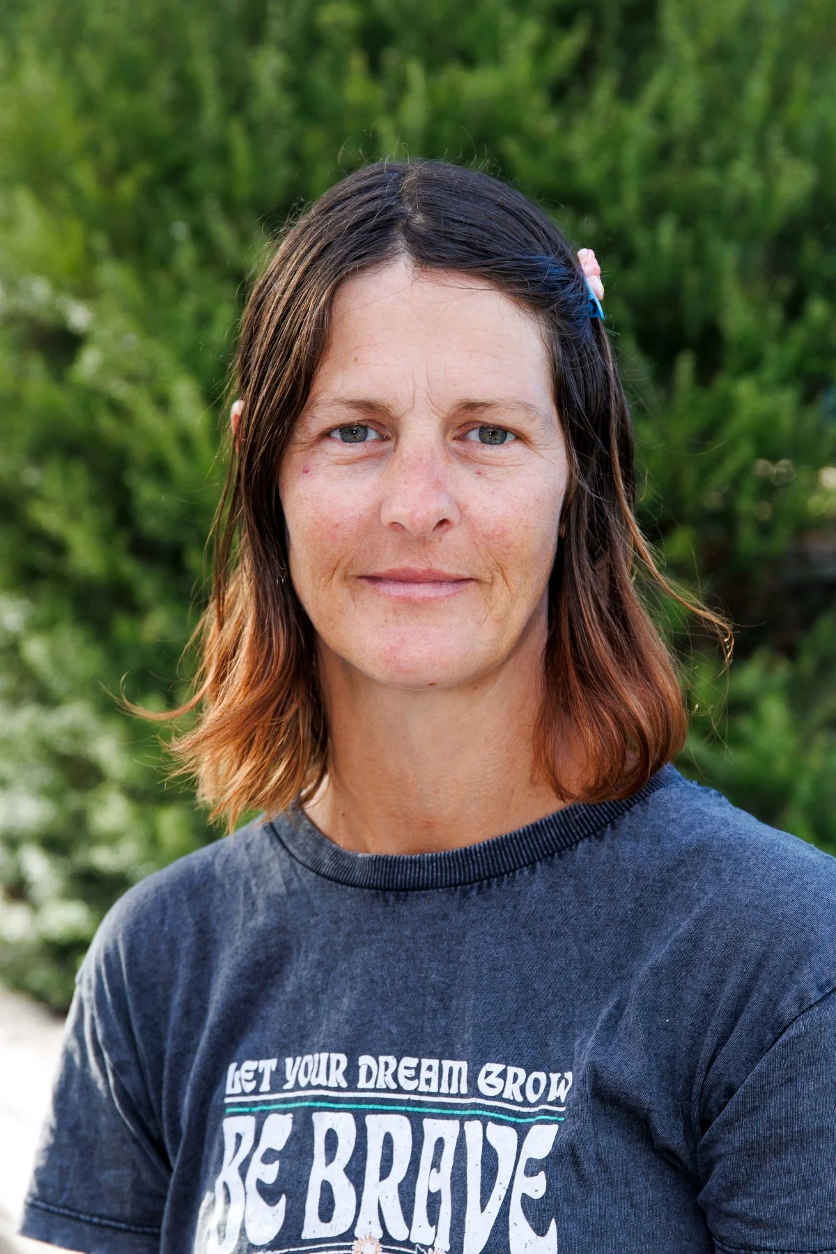 A woman with shoulder-length hair, wearing a dark T-shirt with a motivational quote, standing outdoors with a background of green foliage.