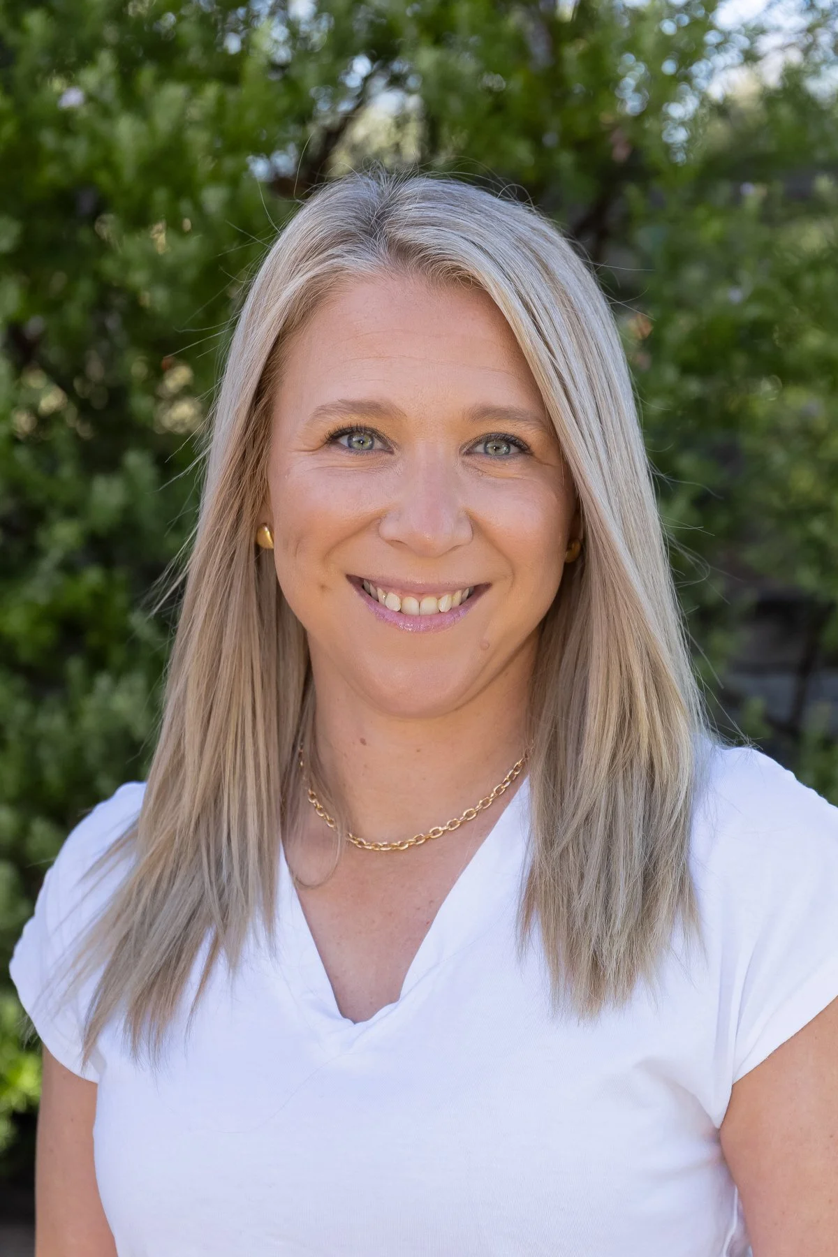 A woman with blonde hair smiling outdoors, wearing a white top, gold earrings, and a gold chain necklace, with trees in the background.