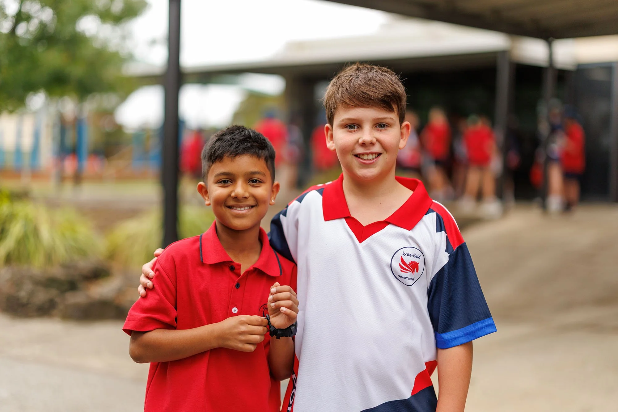 Two schoolboys smiling and standing close together outdoors, one with his arm around the other. Both are wearing school uniforms, one in a red polo shirt and the other in a white, red, and blue sports jersey with a school logo. There is a school playground in the background.