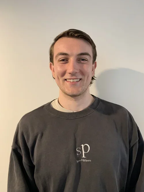 A young man with short brown hair and a light beard, smiling, wearing a gray sweatshirt with a small logo 'sp' and 'SportsMedMeds' underneath, standing against a plain off-white wall.