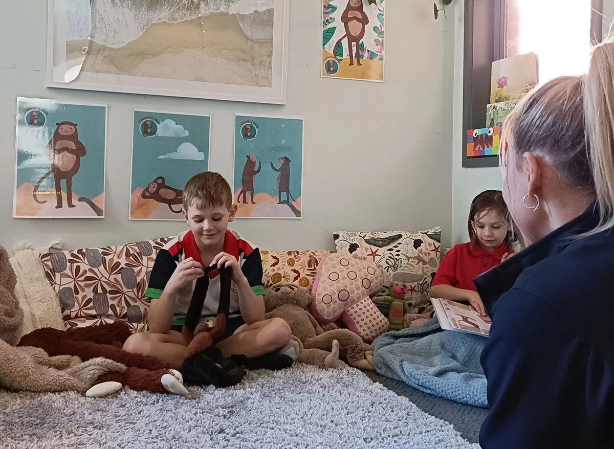 Two children, a boy and a girl, are sitting on a cozy rug in a living room; an adult woman is sitting with them, reading a book. The room has plush toys and decorative pillows, with framed animal-themed artwork on the wall.