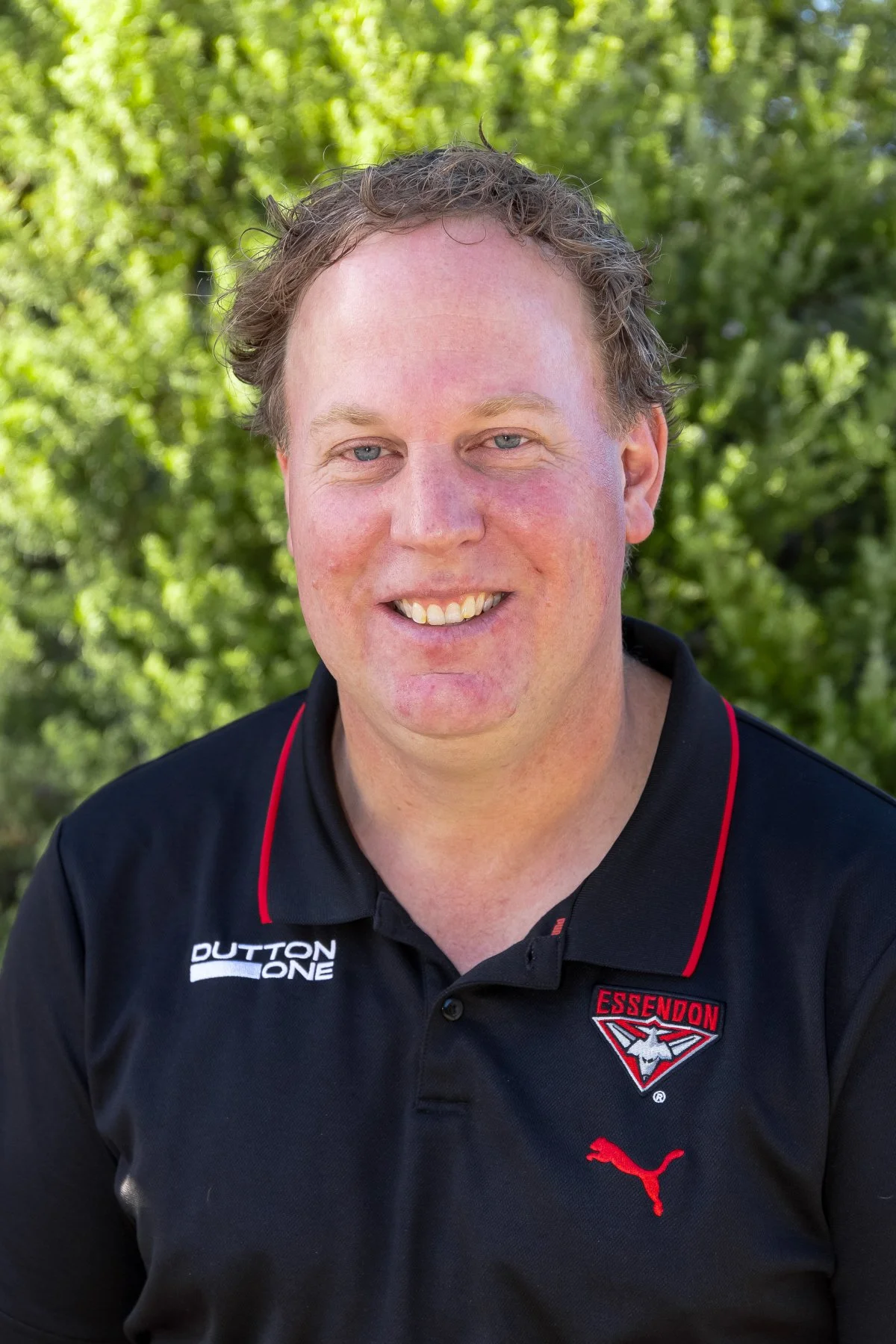 A man with light skin, curly brown hair, and blue eyes smiling outdoors, wearing a black sports shirt with red accents, a Puma logo, and the Essendon football club crest.