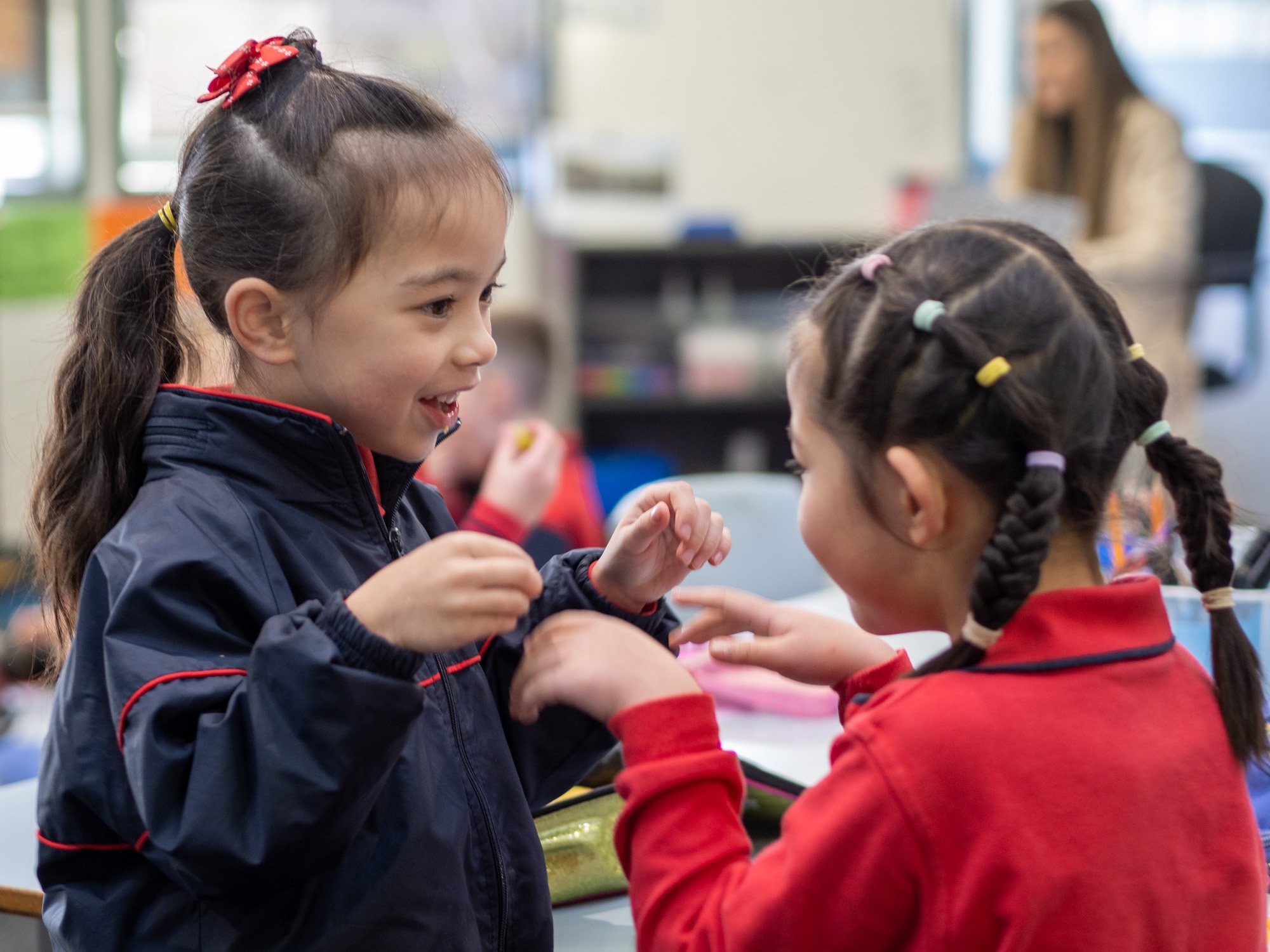 Two young girls with braided hair and colorful hair ties are smiling and interacting with each other in a classroom setting, with a teacher and other children visible in the background.