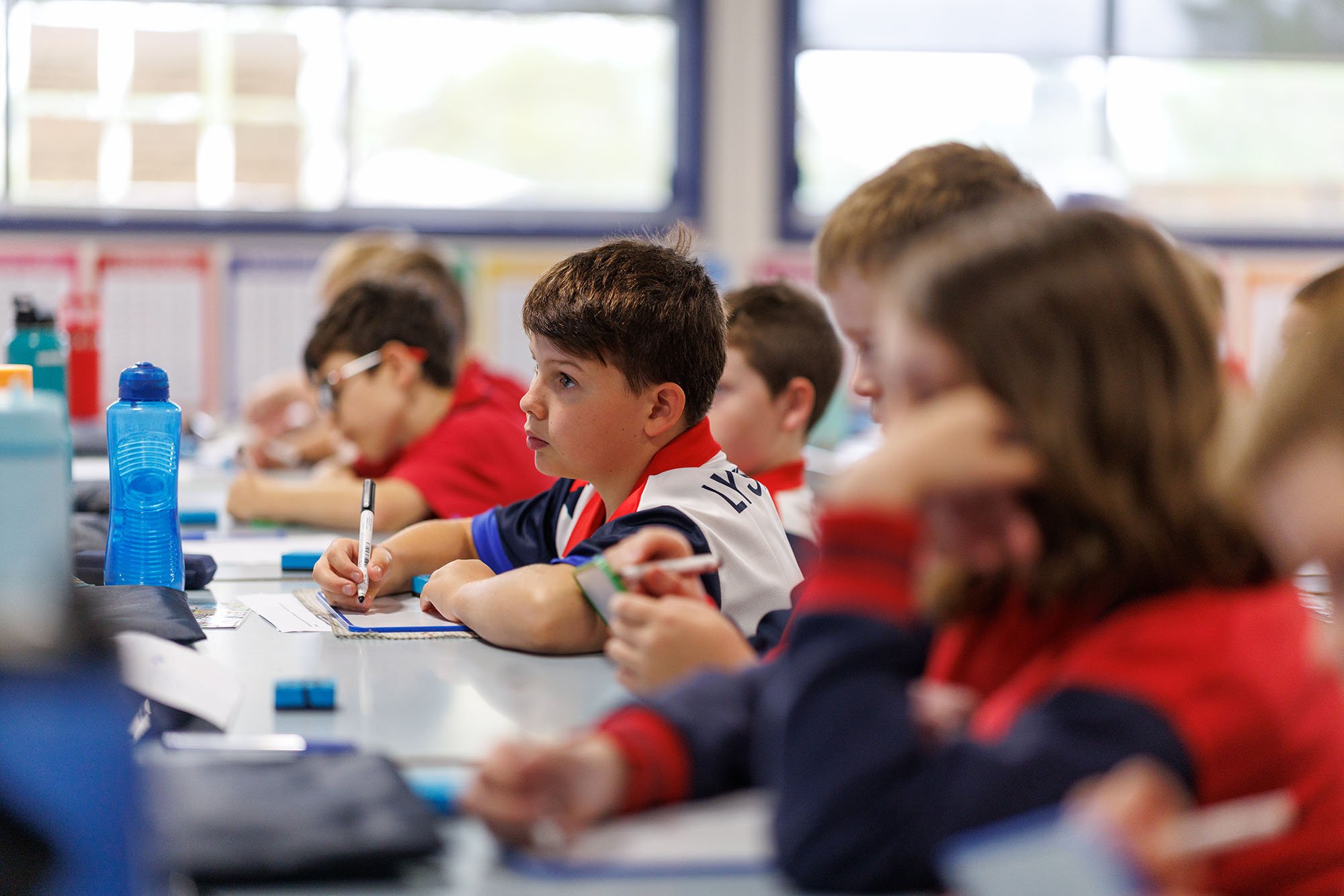 School children sitting at desks in a classroom, attentively listening and taking notes, with water bottles and school supplies on the desks.