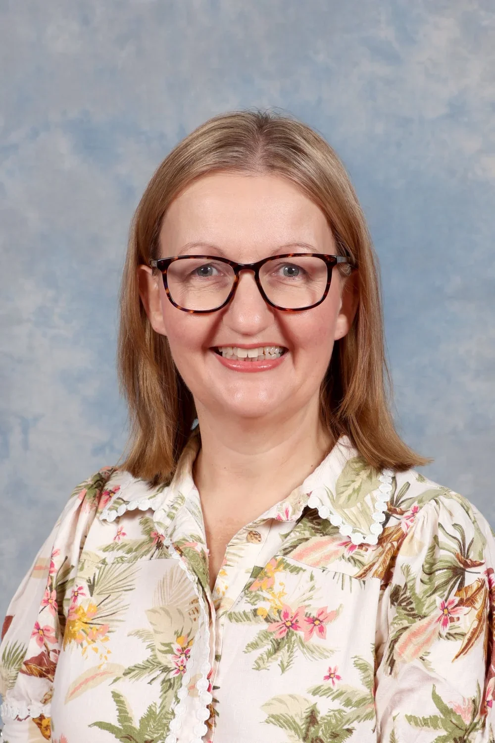 A woman with shoulder-length light brown hair, wearing glasses with a tortoise shell pattern, a floral blouse, and smiling, against a soft blue background.