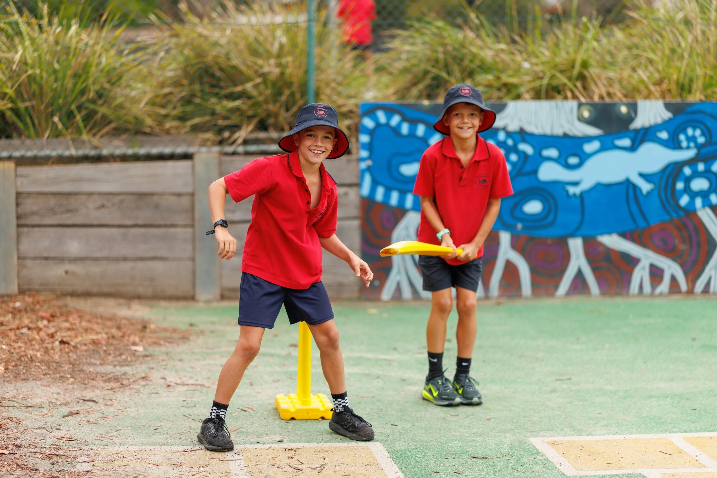 Two children wearing red shirts, blue shorts, and hats playing with a yellow bat and ball outside near a painted mural.