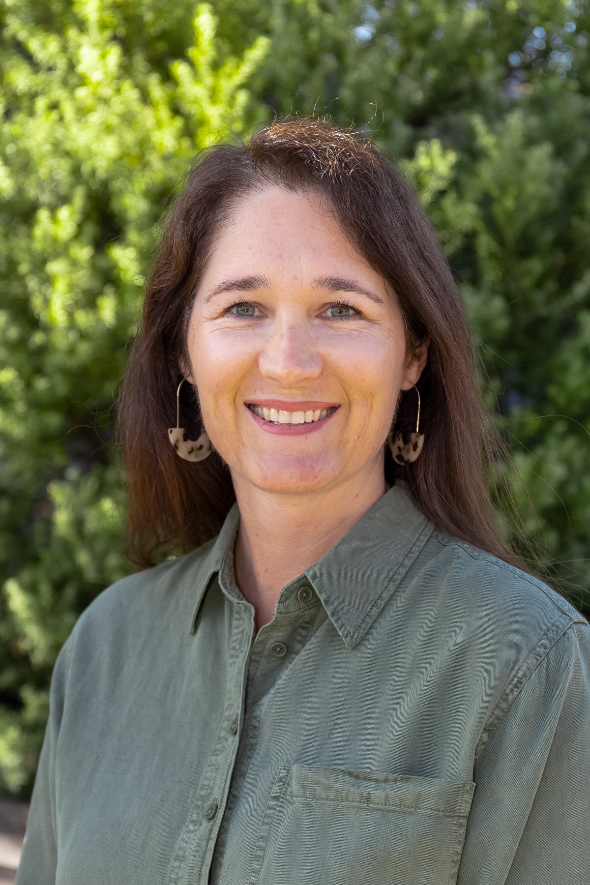 A woman with long brown hair, wearing hoop earrings and a green button-up shirt, smiling outdoors with green trees in the background.