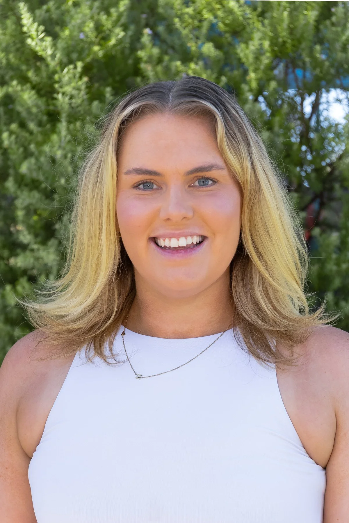 A young woman with blonde hair, blue eyes, and a bright smile, wearing a white sleeveless top and a delicate necklace, standing outdoors with green trees in the background.