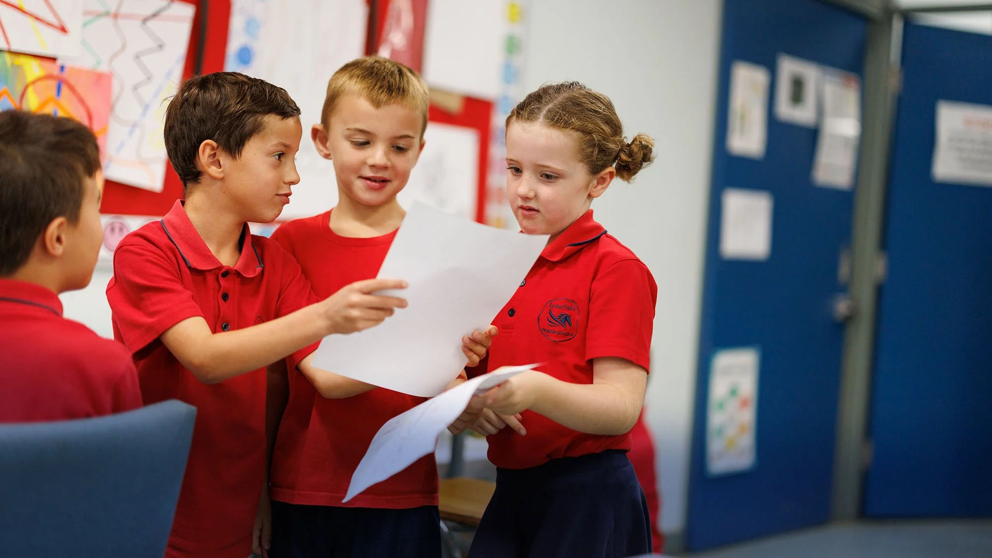 Four children in red school uniforms stand together in a classroom, looking at papers and discussing.