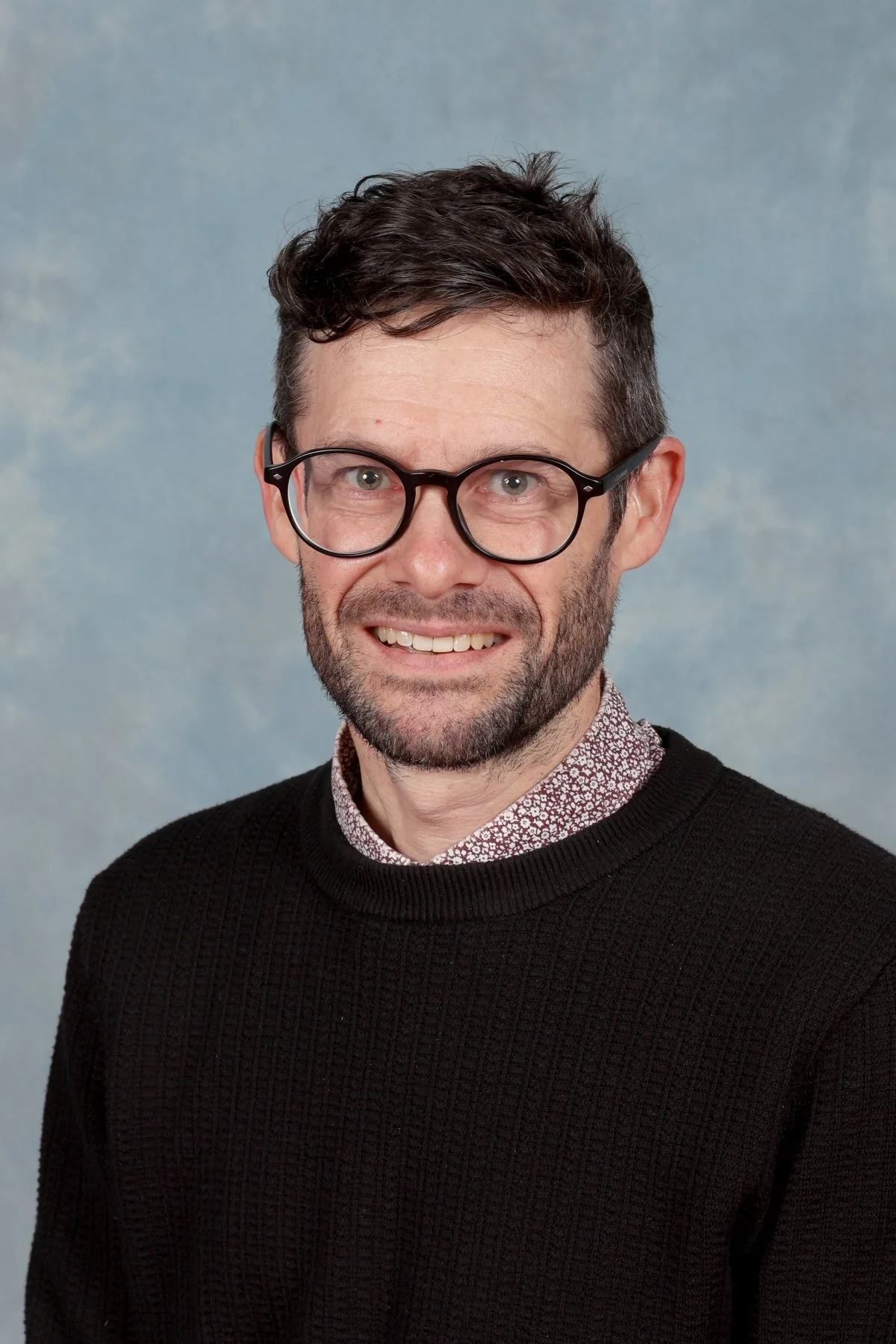 A man with dark hair, glasses, and a beard, smiling in front of a light blue background.