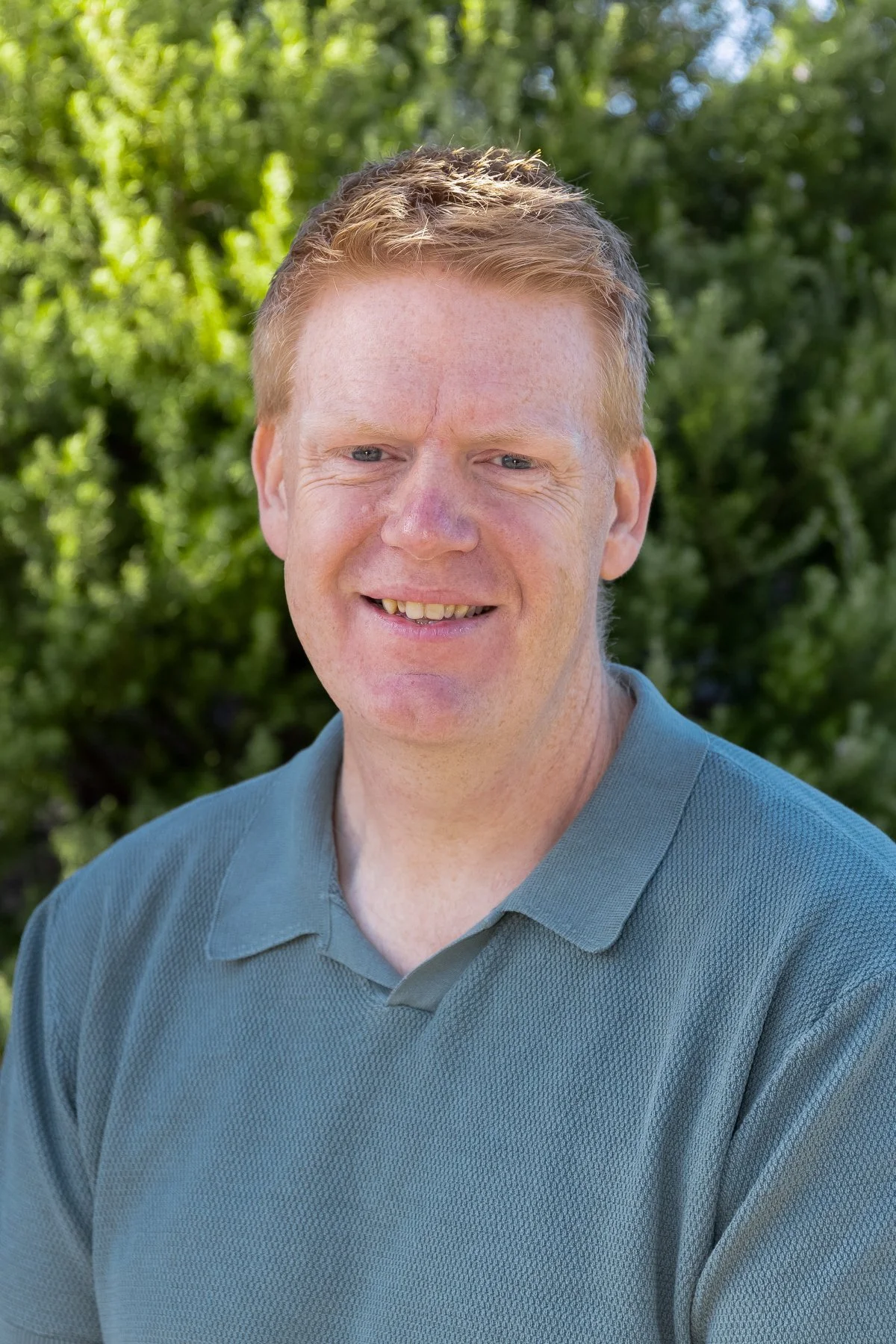 A smiling man with short, light brown hair and freckles, wearing a green polo shirt, standing outdoors in front of green trees.