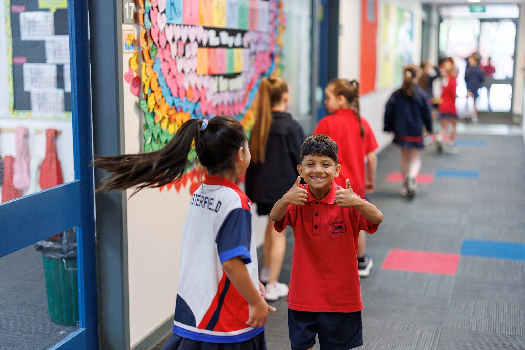 Smiling boy giving two thumbs up in a school hallway with other children in the background.