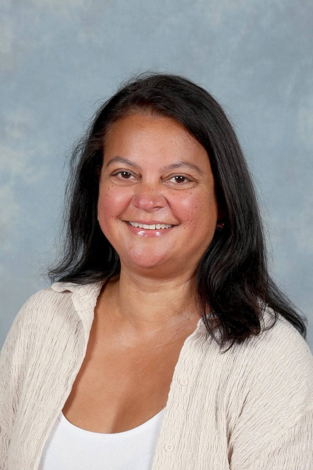 A smiling woman with black hair wearing a cream-colored cardigan over a white top, posed against a light blue and gray backdrop.