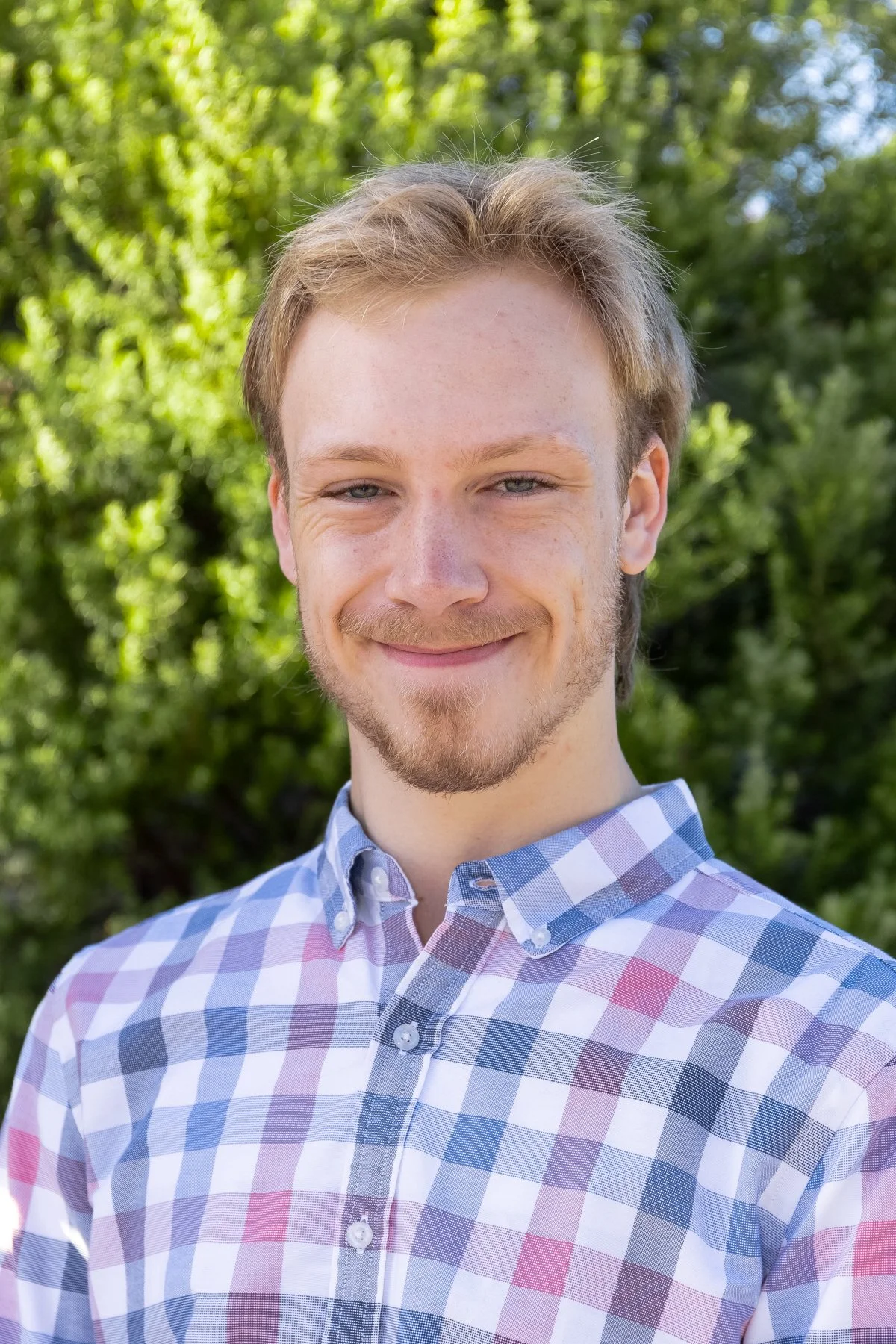 Portrait of a young man smiling outdoors, wearing a checkered shirt, with green trees in the background.