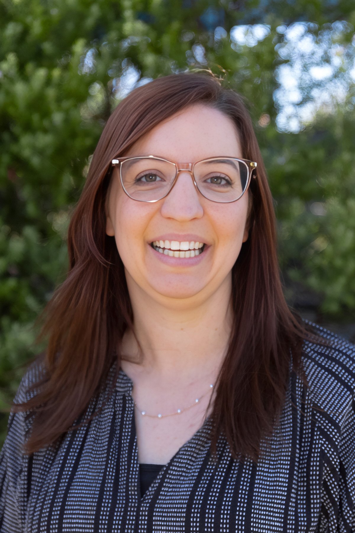 A woman with brown hair and glasses smiling outdoors with greenery in the background.