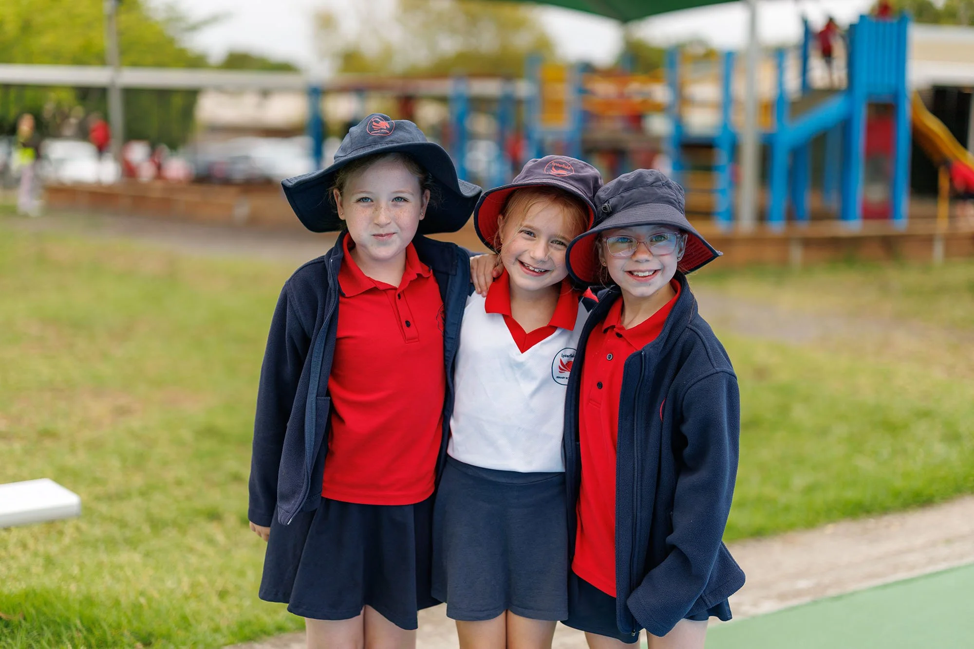 Three young girls in school uniforms and hats standing outdoors at a playground, smiling and posing for the camera.