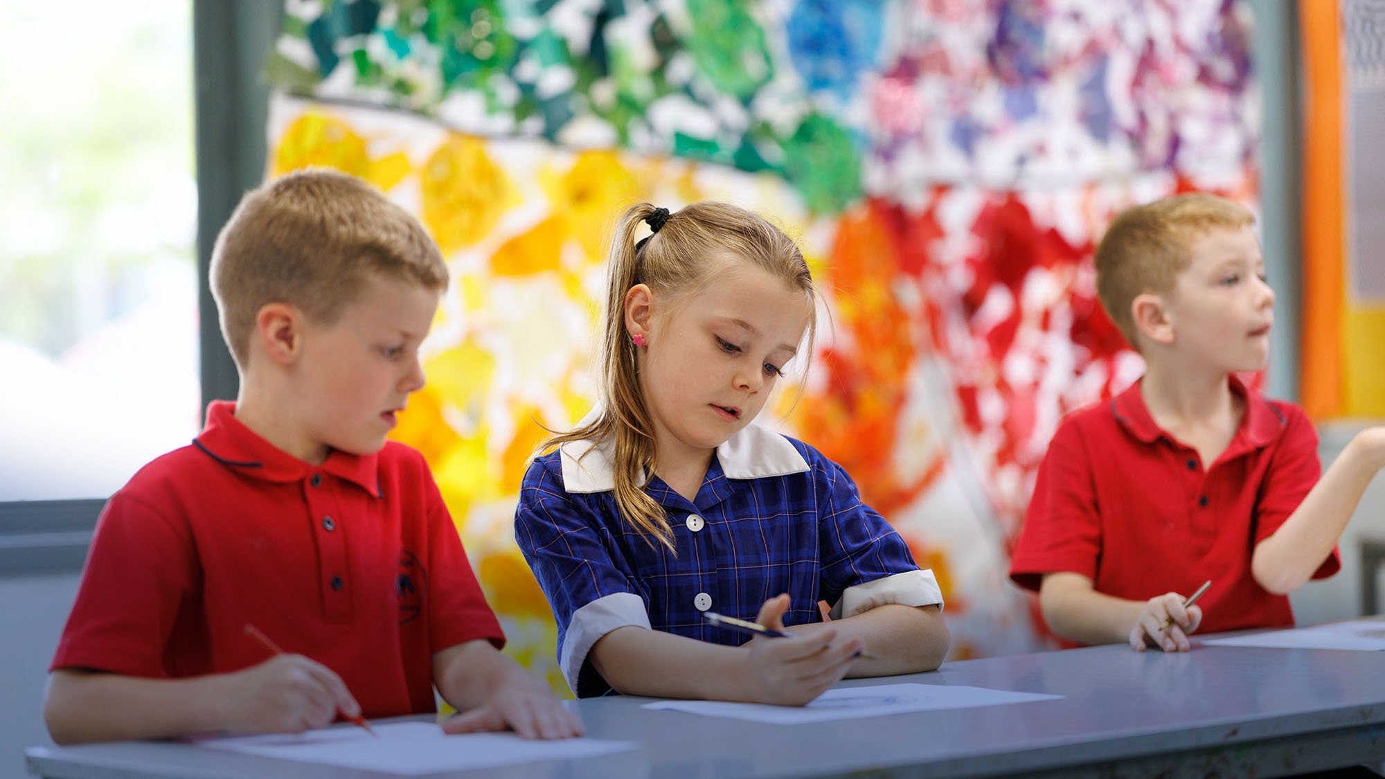 Three children sitting at a table in a classroom, working on papers with pencils, with colorful abstract art on the wall behind them.
