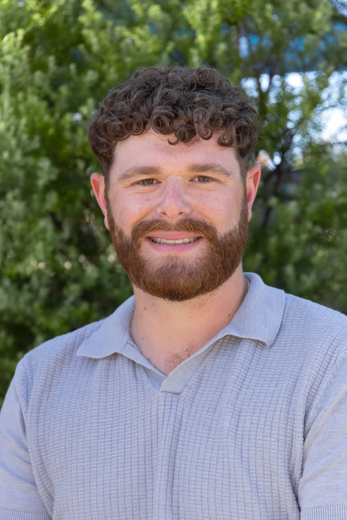 A smiling man with curly brown hair and a beard outdoors with green trees in the background.