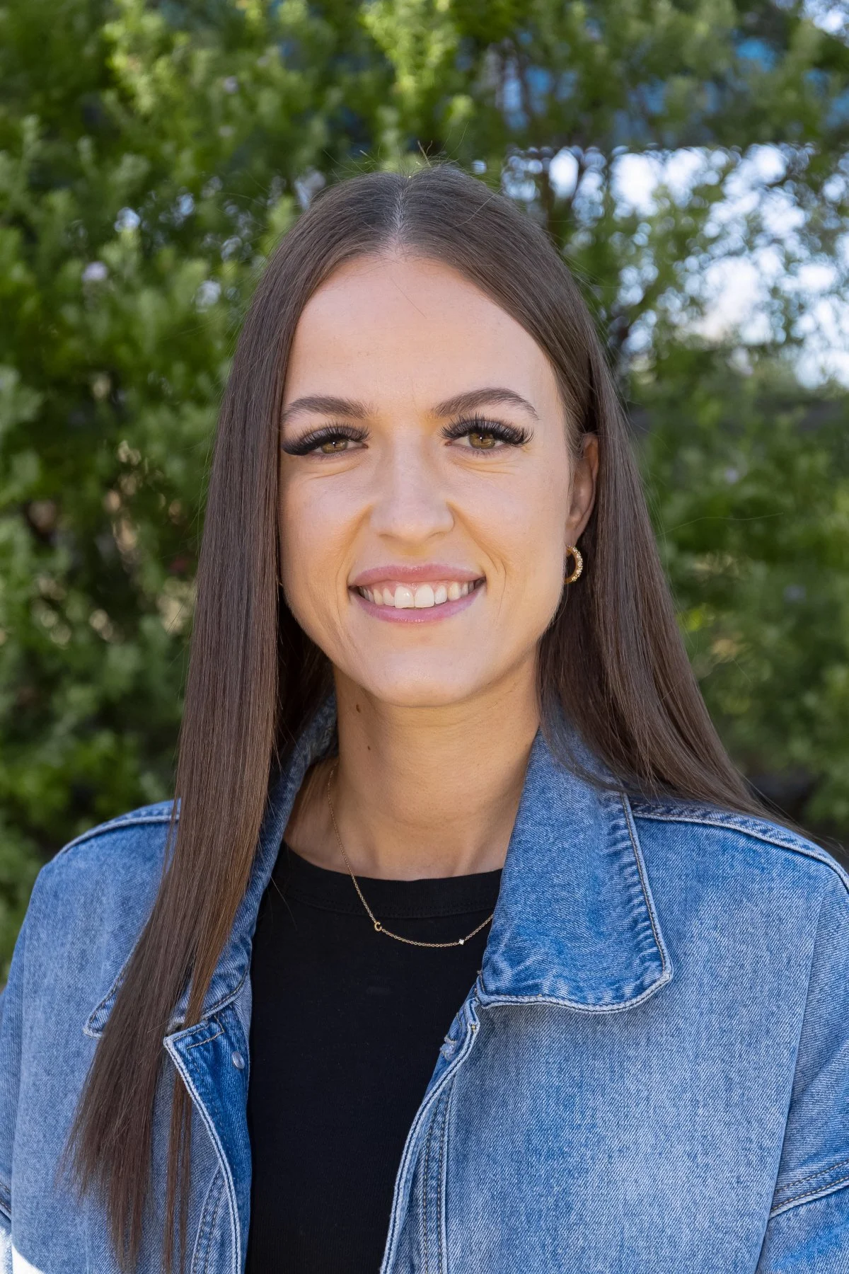 A young woman with long brown hair, wearing a denim jacket and black top, smiling outdoors with green trees in the background.