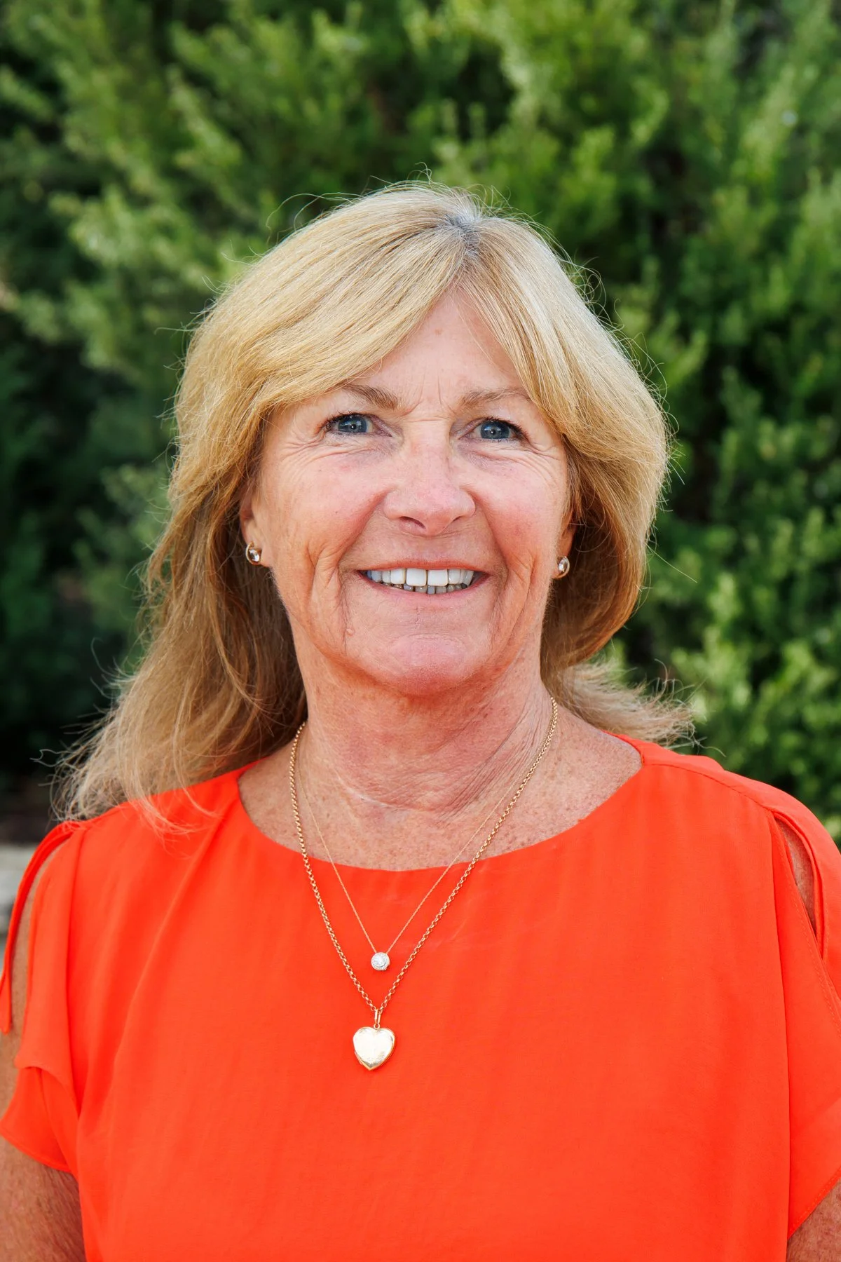 A smiling middle-aged woman with blond hair and blue eyes, wearing a bright orange top and layered gold necklaces, stands outdoors with green foliage in the background.