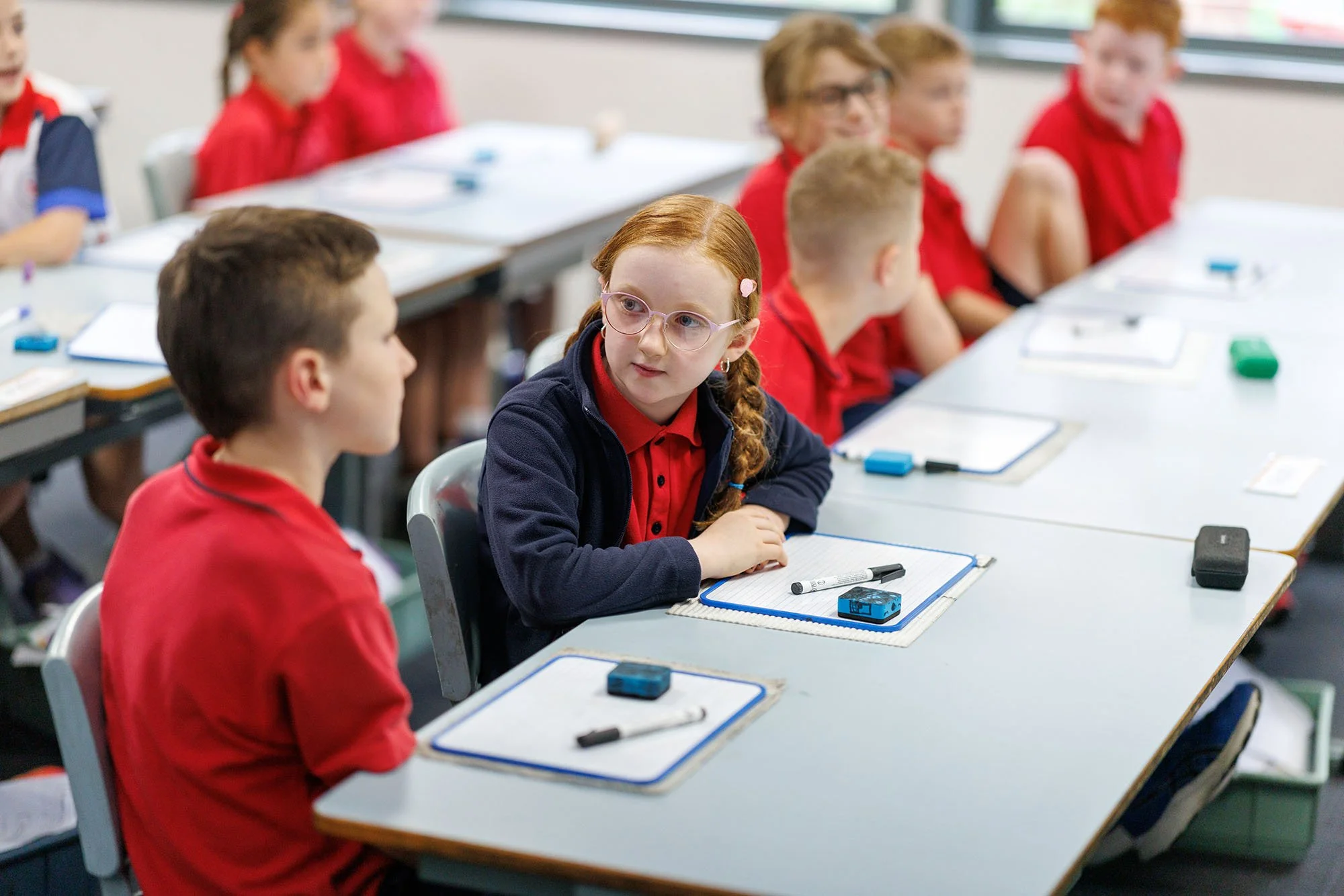 A classroom with children wearing red uniforms, one girl with glasses and a braid talking to a boy, sitting at desks with notebooks and writing supplies.